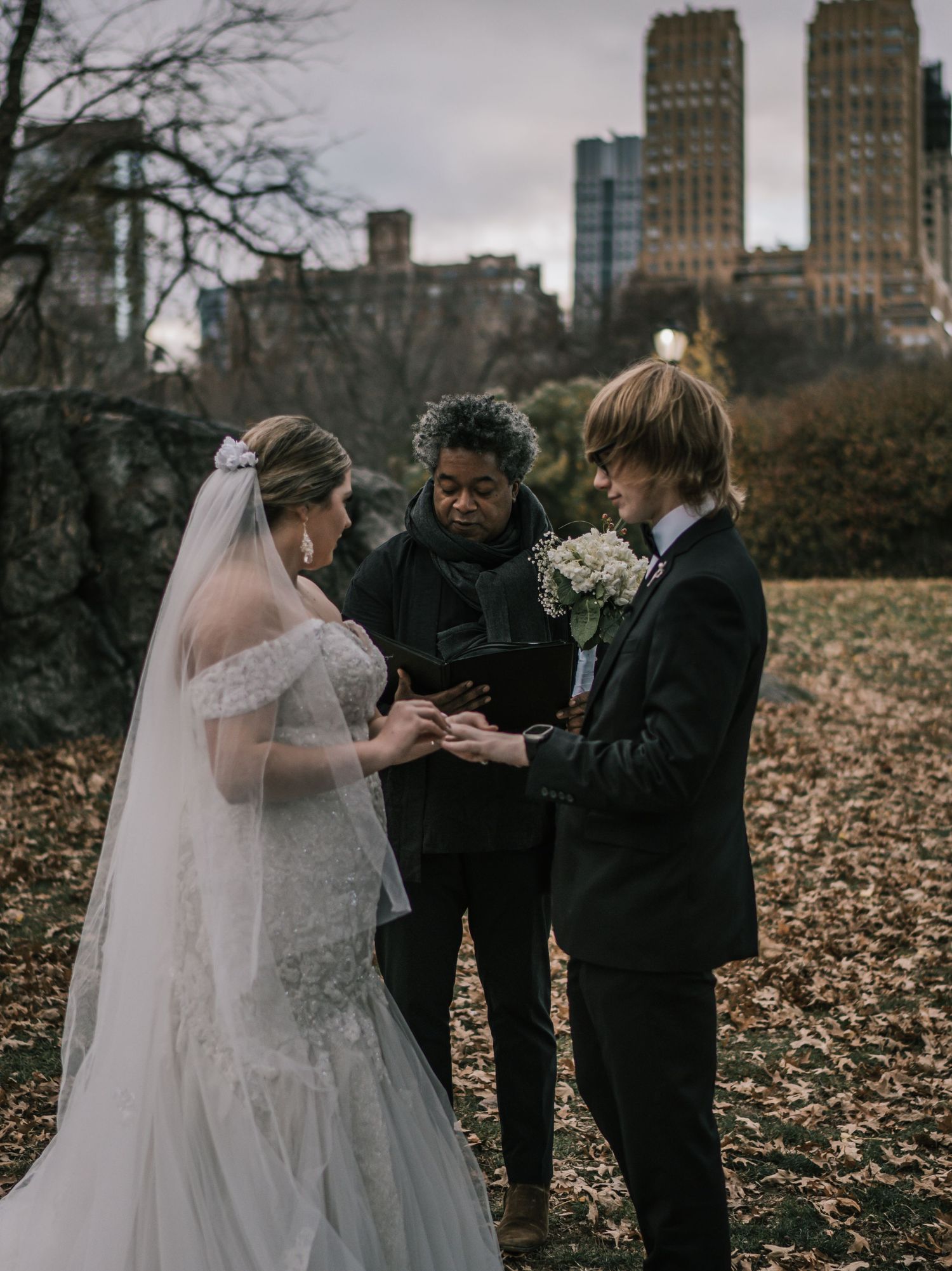 Couple exchanges rings during intimate Central Park elopement ceremony at dusk with city buildings in background.