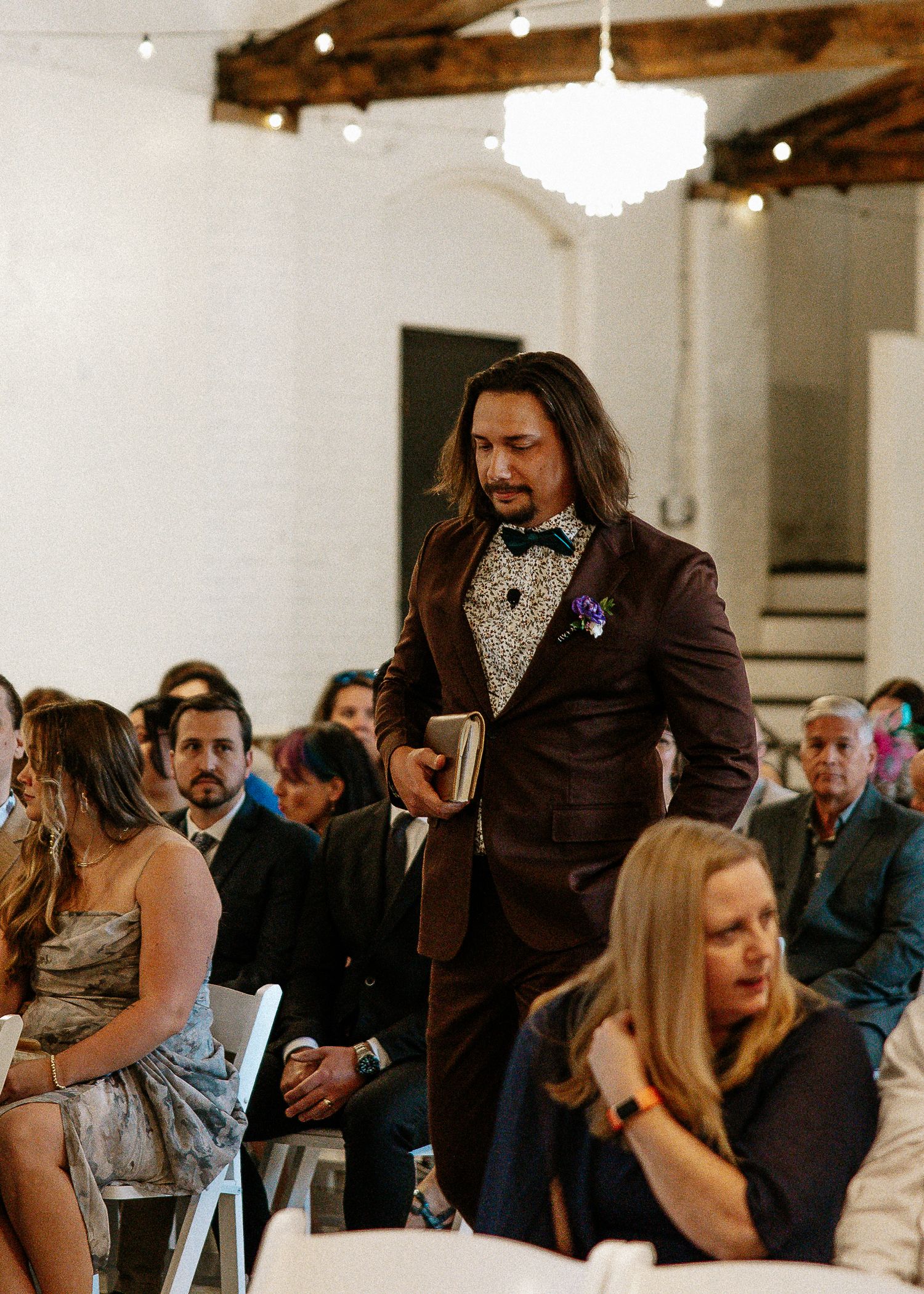Man in brown suit with floral shirt and bow tie walking down wedding ceremony aisle holding book.