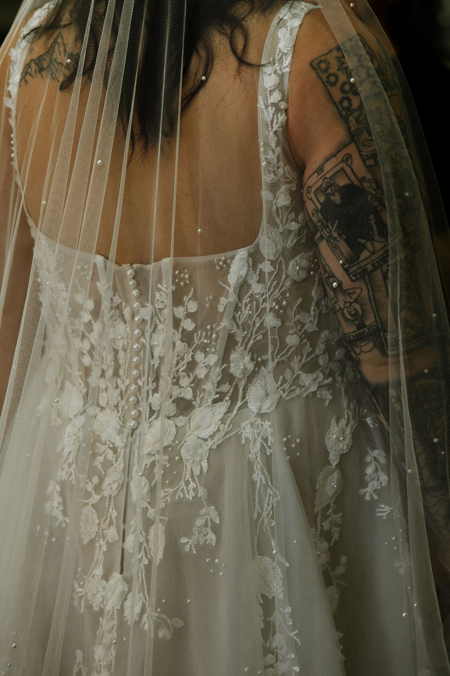 Close-up of tattooed bride in intricate floral lace wedding gown with pearl-embellished veil from behind.
