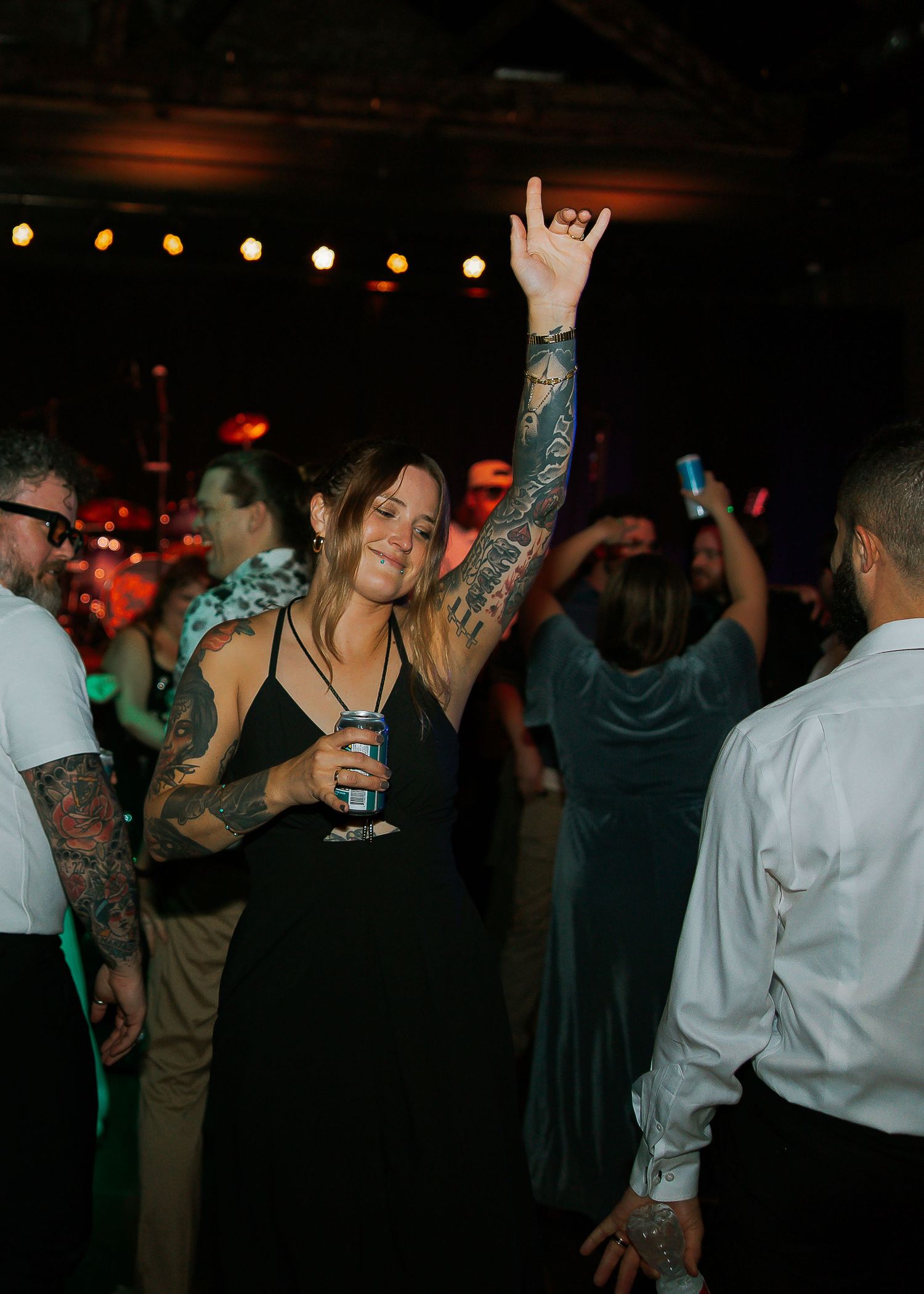 Tattooed woman in black dress raising hand with rock sign on a crowded dance floor.
