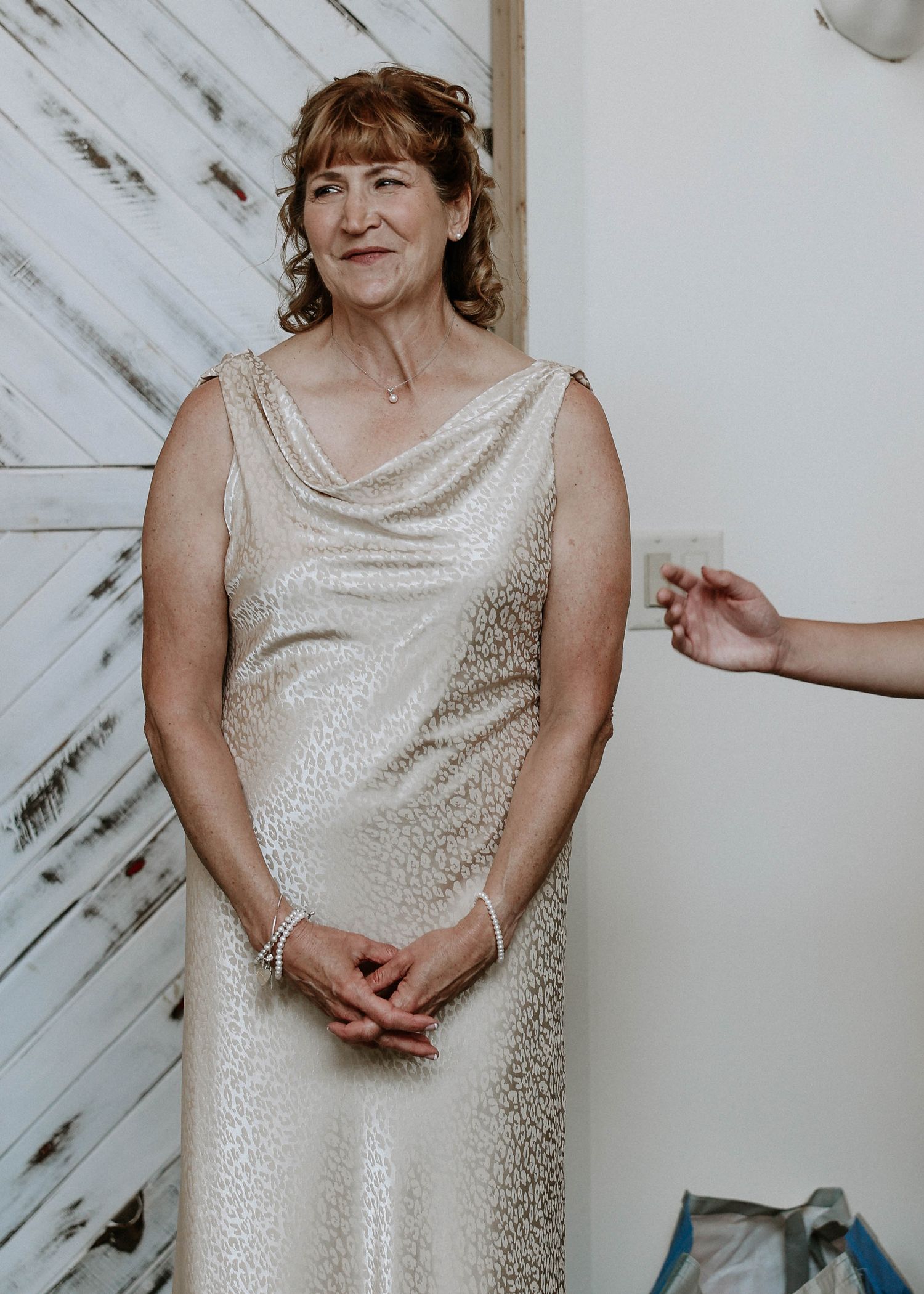Older woman in elegant sleeveless gold gown standing before white barn wood backdrop.