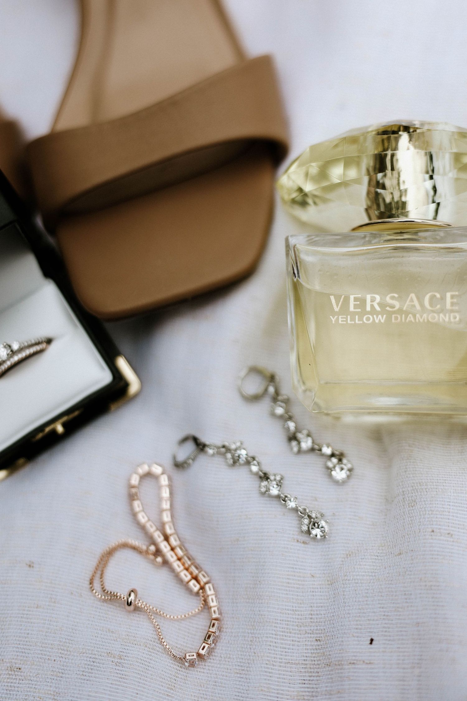 Versace Yellow Diamond perfume displayed with tan sandals, diamond earrings, and delicate rose gold bracelet on white fabric.