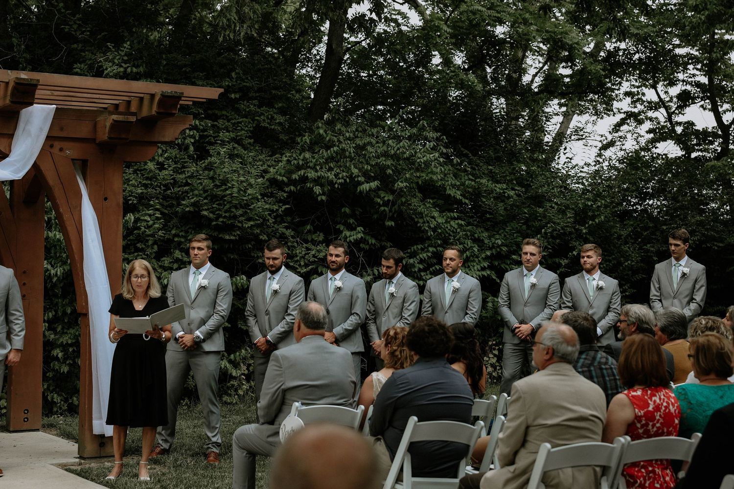 Groomsmen in gray suits stand at outdoor wedding ceremony beneath wooden arbor surrounded by lush green trees.