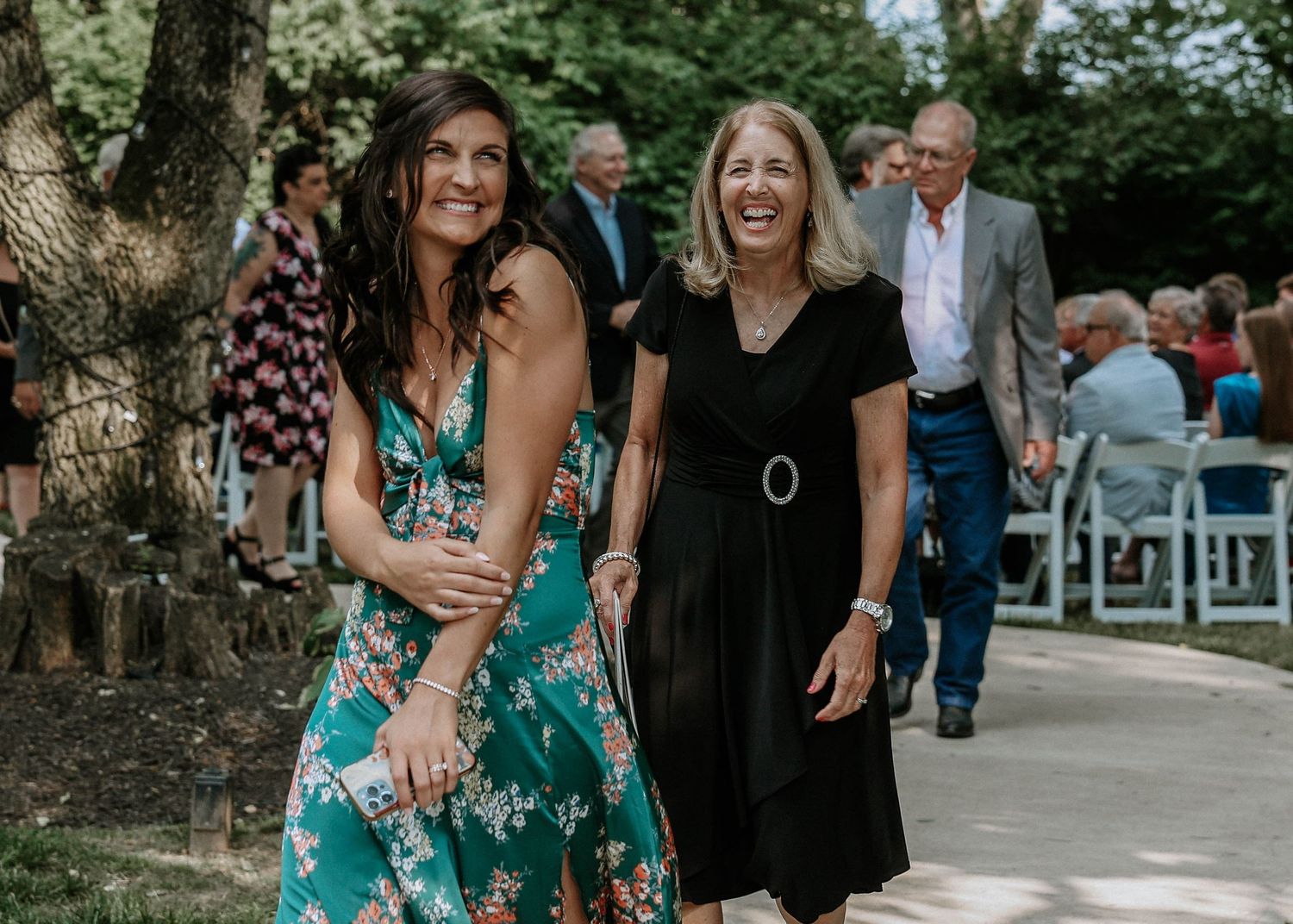 Two women smiling and walking together at an outdoor wedding ceremony.