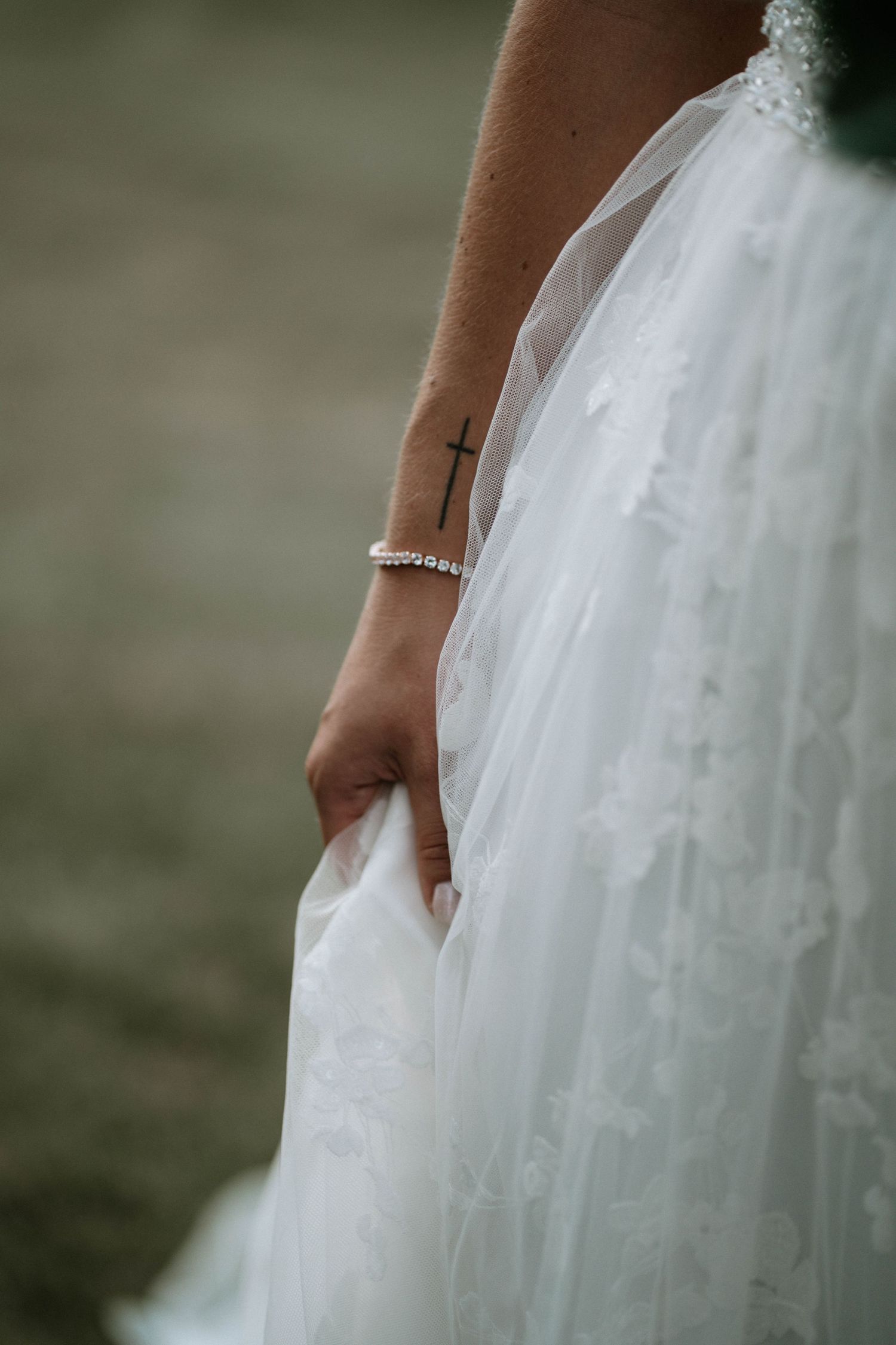 Bride holding white lace wedding dress with cross tattoo and diamond bracelet.