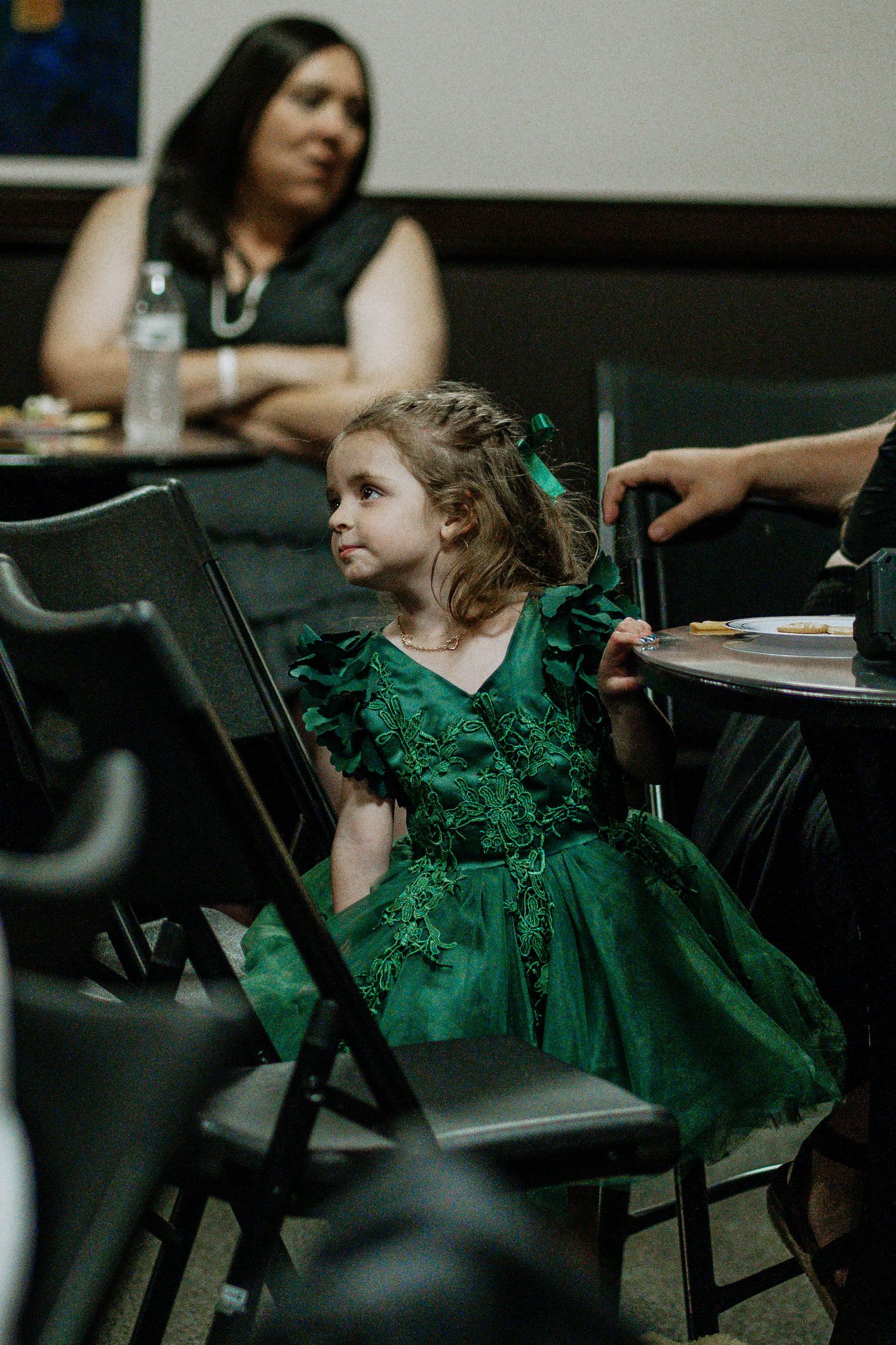 Young girl in emerald green ruffled dress sits at table during formal event, woman in black dress seated behind her.