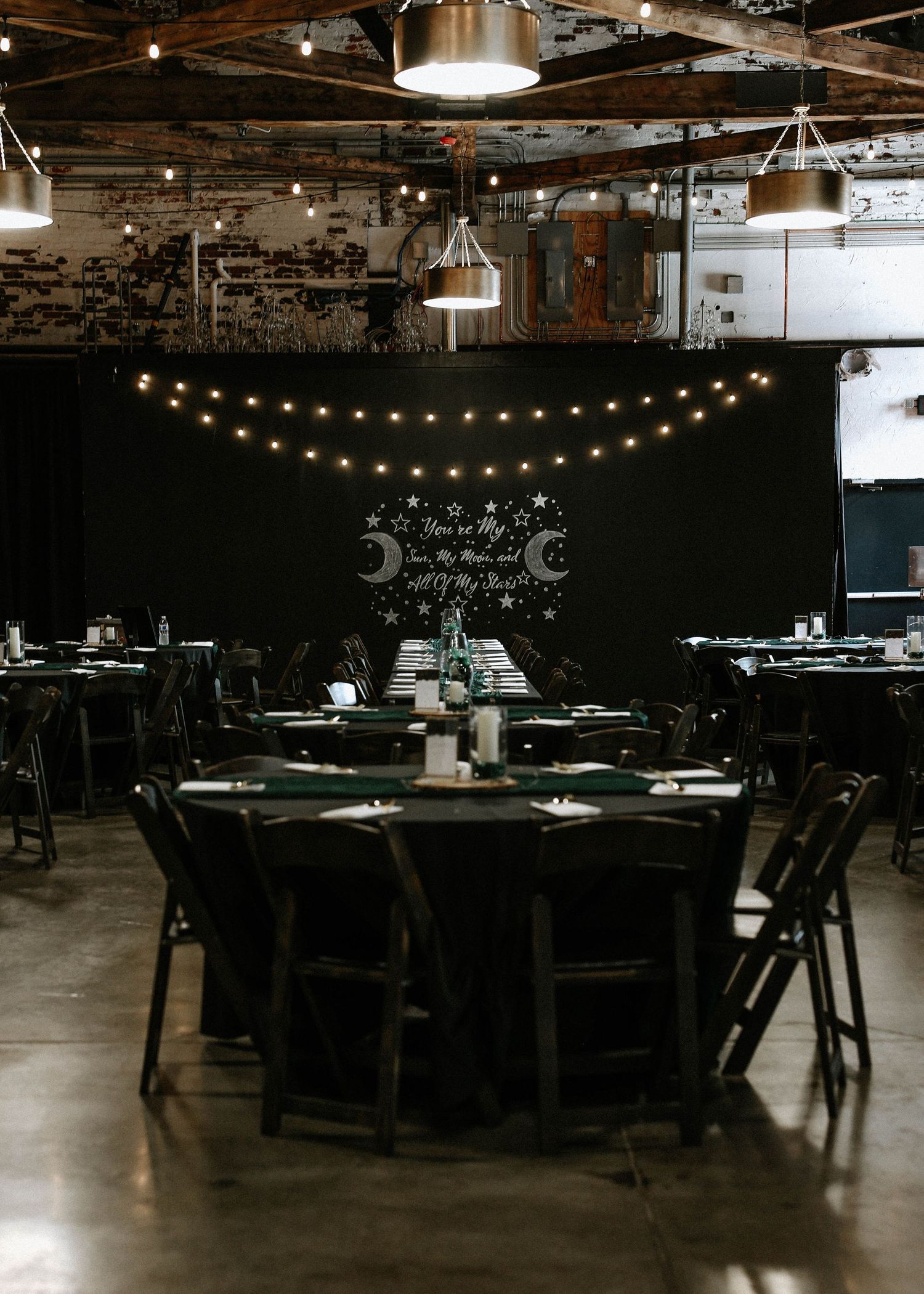 Elegant wedding reception with black tables, string lights, and celestial moon backdrop in an industrial venue.