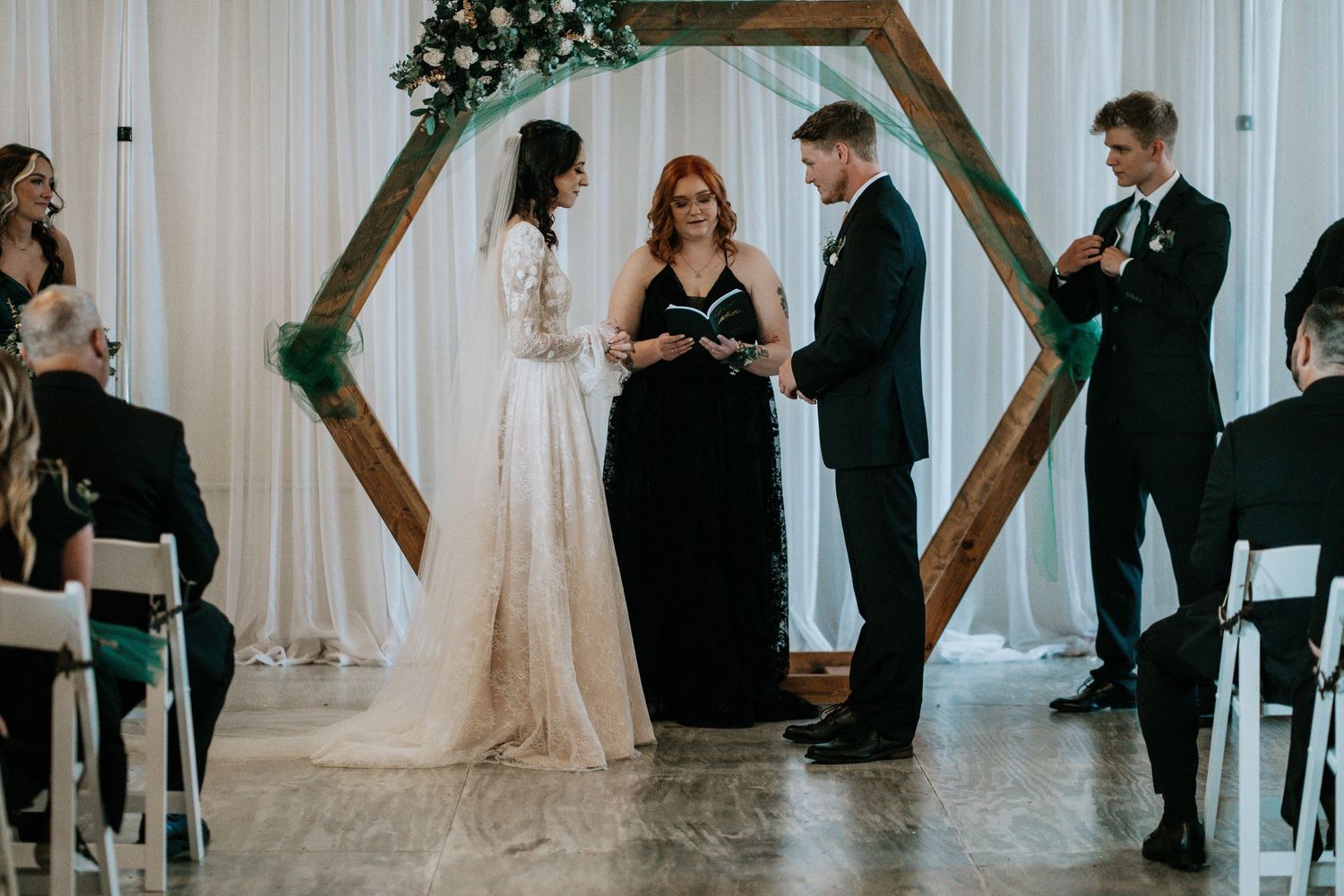 Bride and groom exchange vows at hexagonal wooden arch altar with officiant during elegant indoor wedding ceremony.