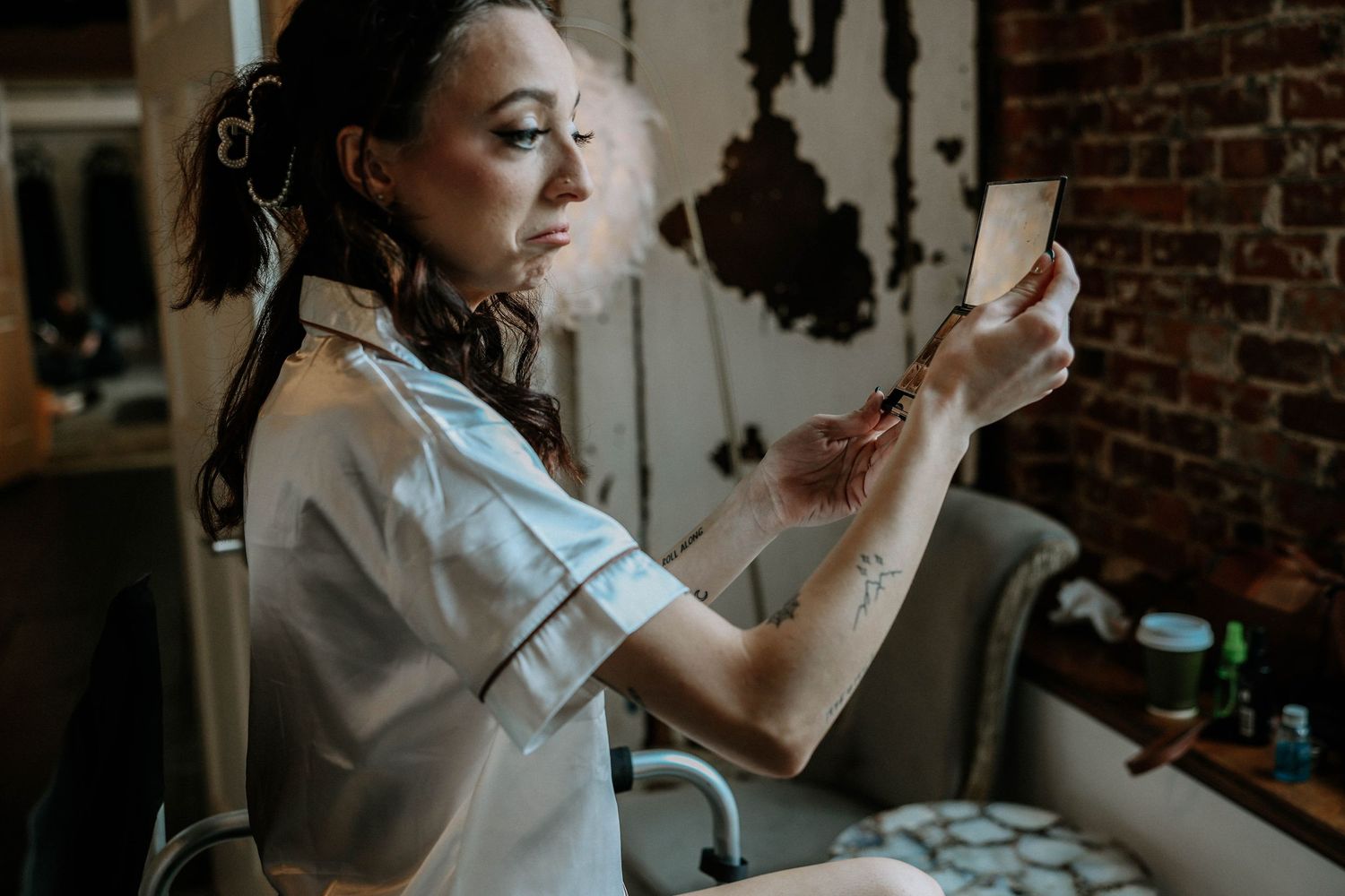 A tattooed woman with dreadlocks in a white shirt examines a small card or photo in a rustic brick-walled studio.