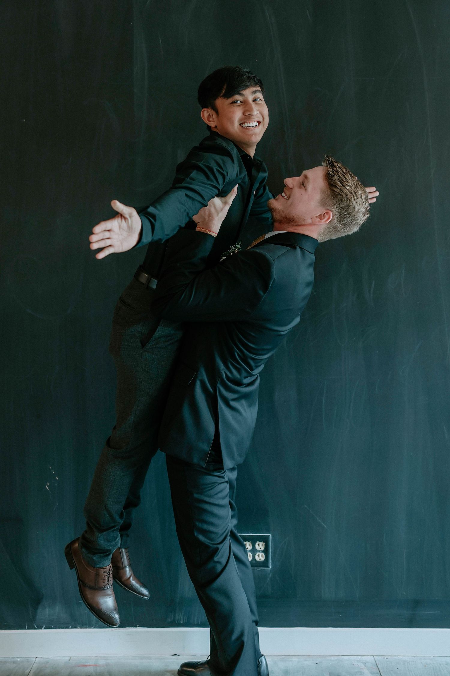 Two men in dark suits playfully pose, one lifting the other in front of a chalkboard wall.