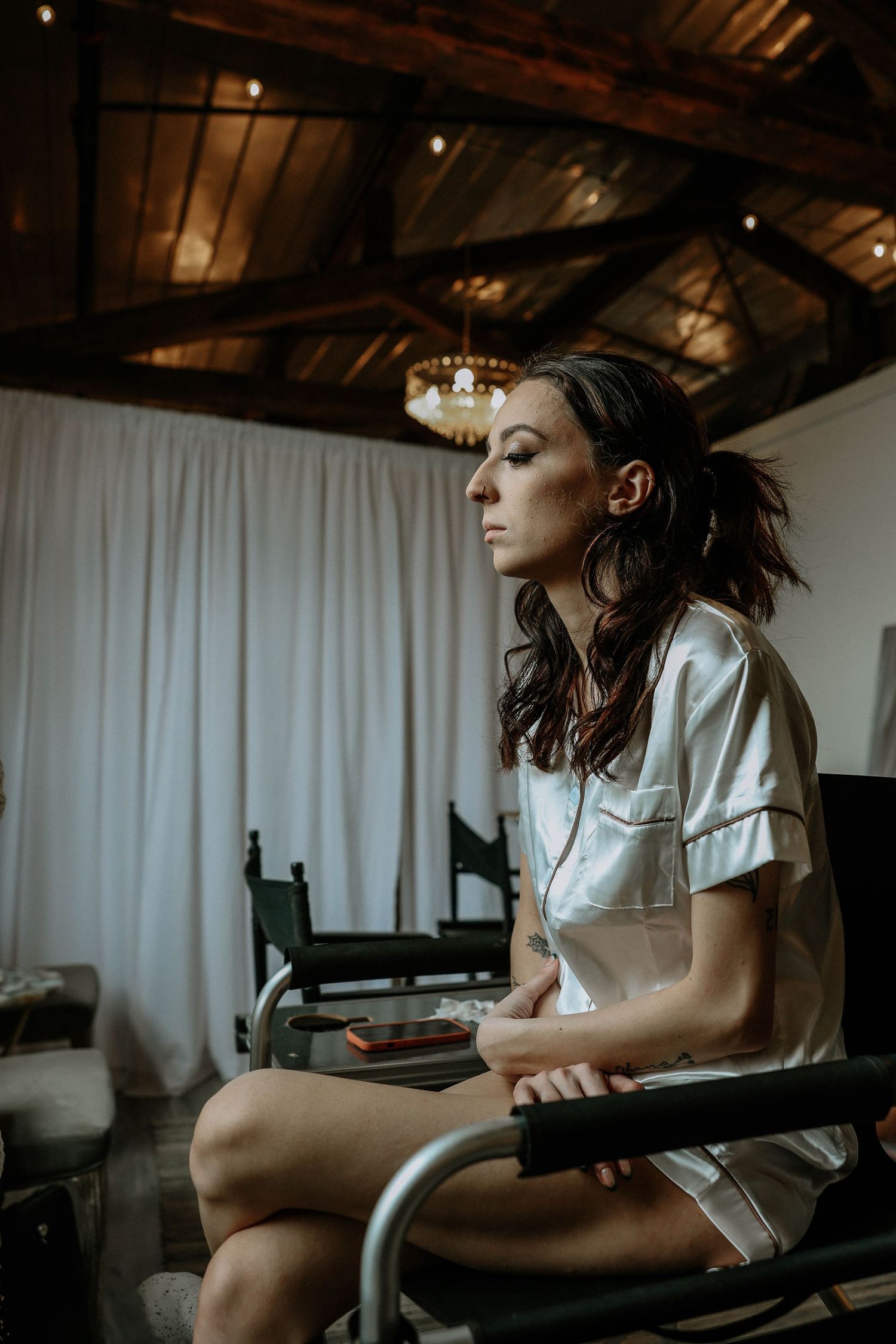 Woman in white shirt sits pensively in a chair inside a rustic room with wooden ceiling beams and chandelier.