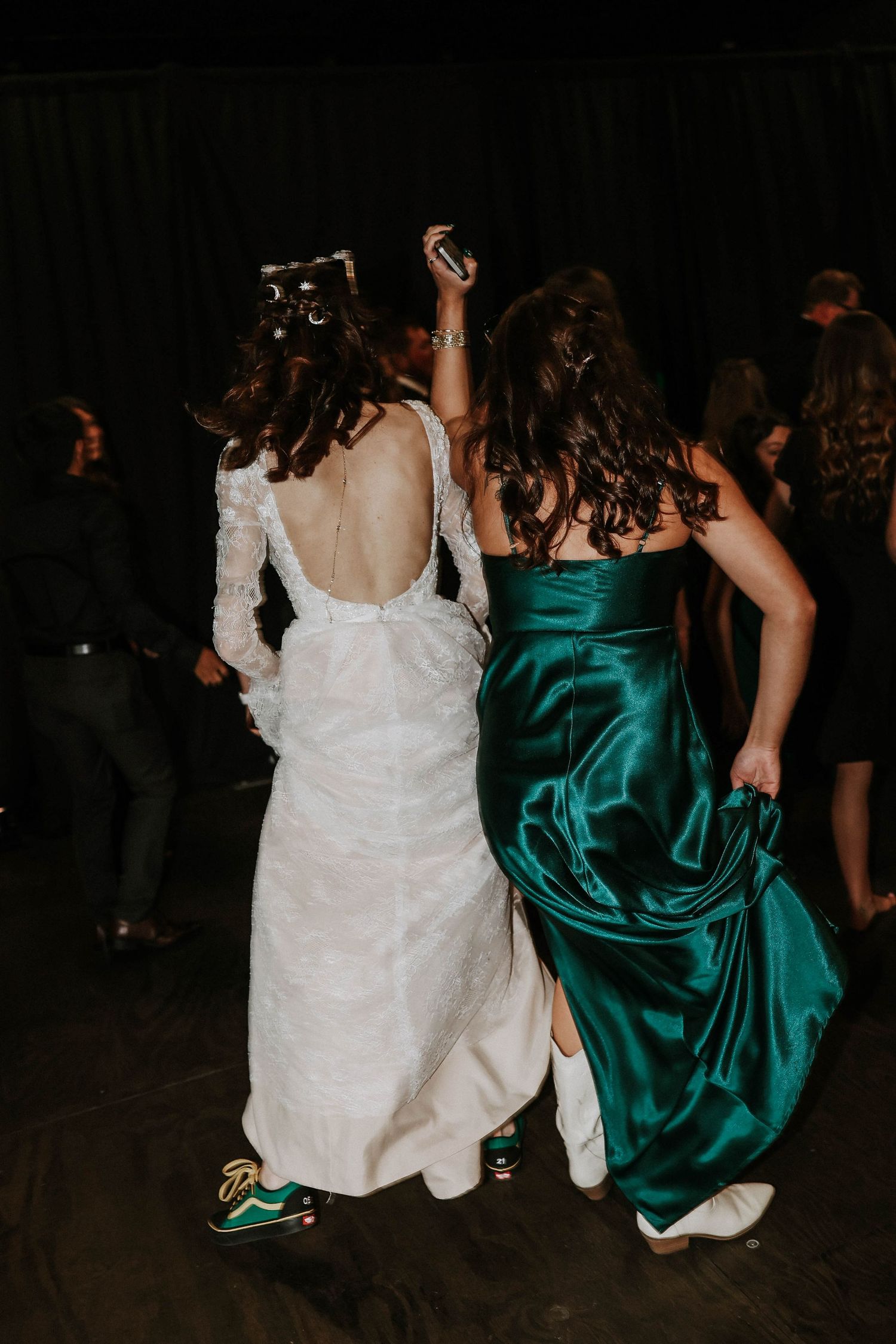 Bride in white lace gown and friend in emerald satin dress dancing together at a wedding reception.
