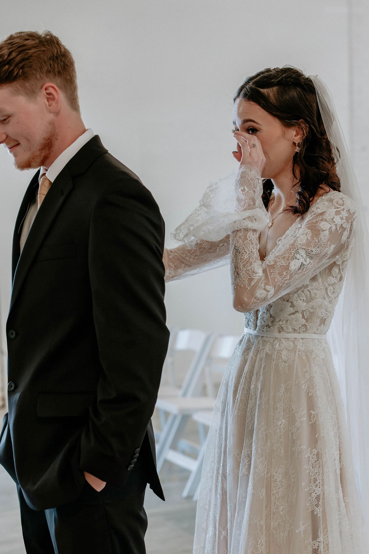 Emotional bride in lace gown wiping tears beside groom in black suit during wedding first look ceremony.