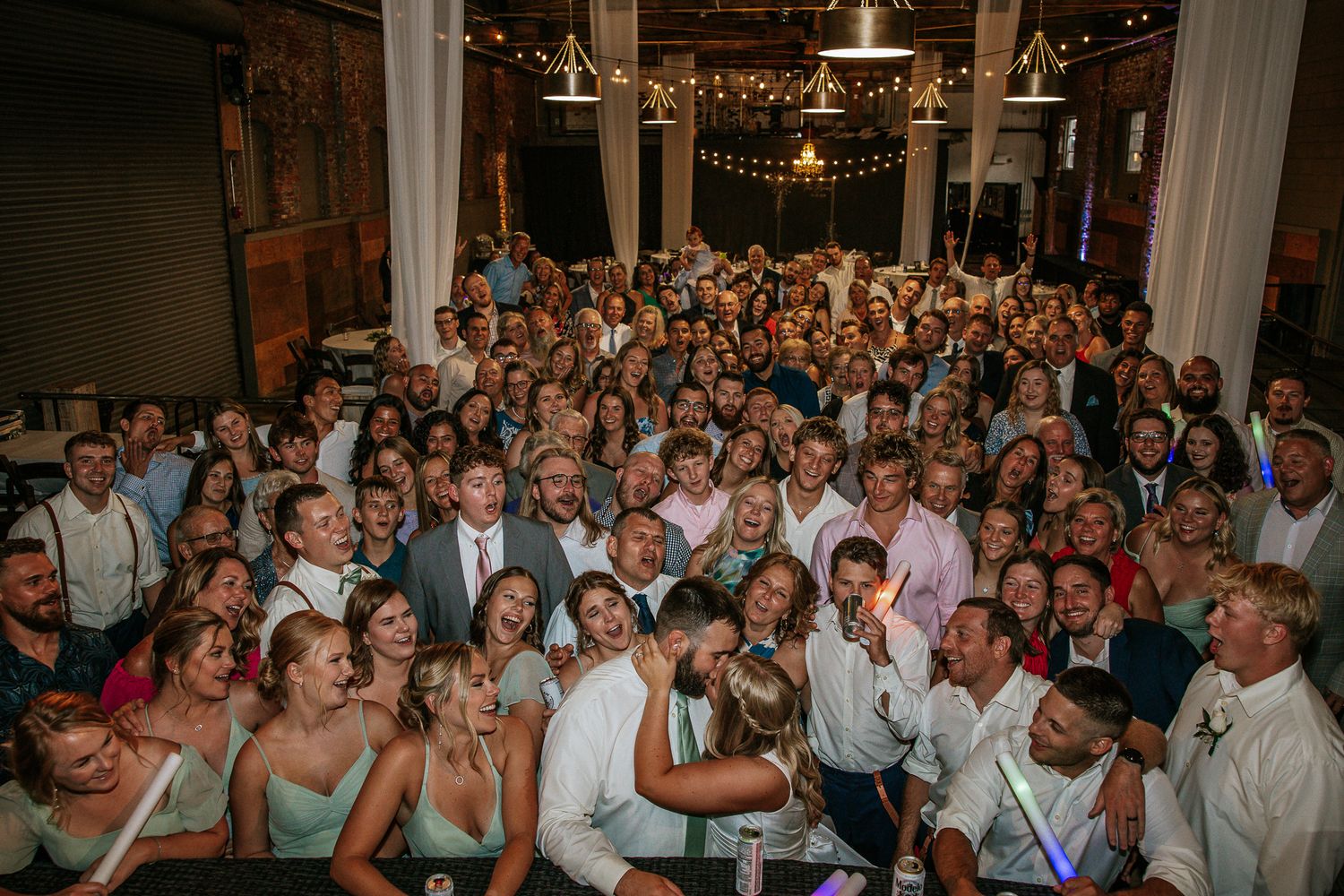 Large wedding reception group photo with bride and groom surrounded by smiling guests indoors.