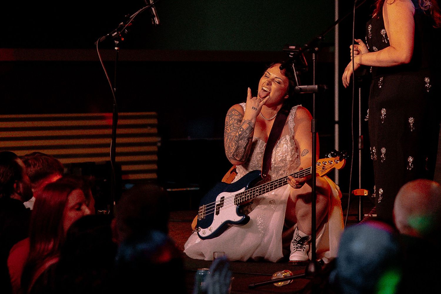 Tattooed bassist in white dress sits on stage playing bass guitar at live show.