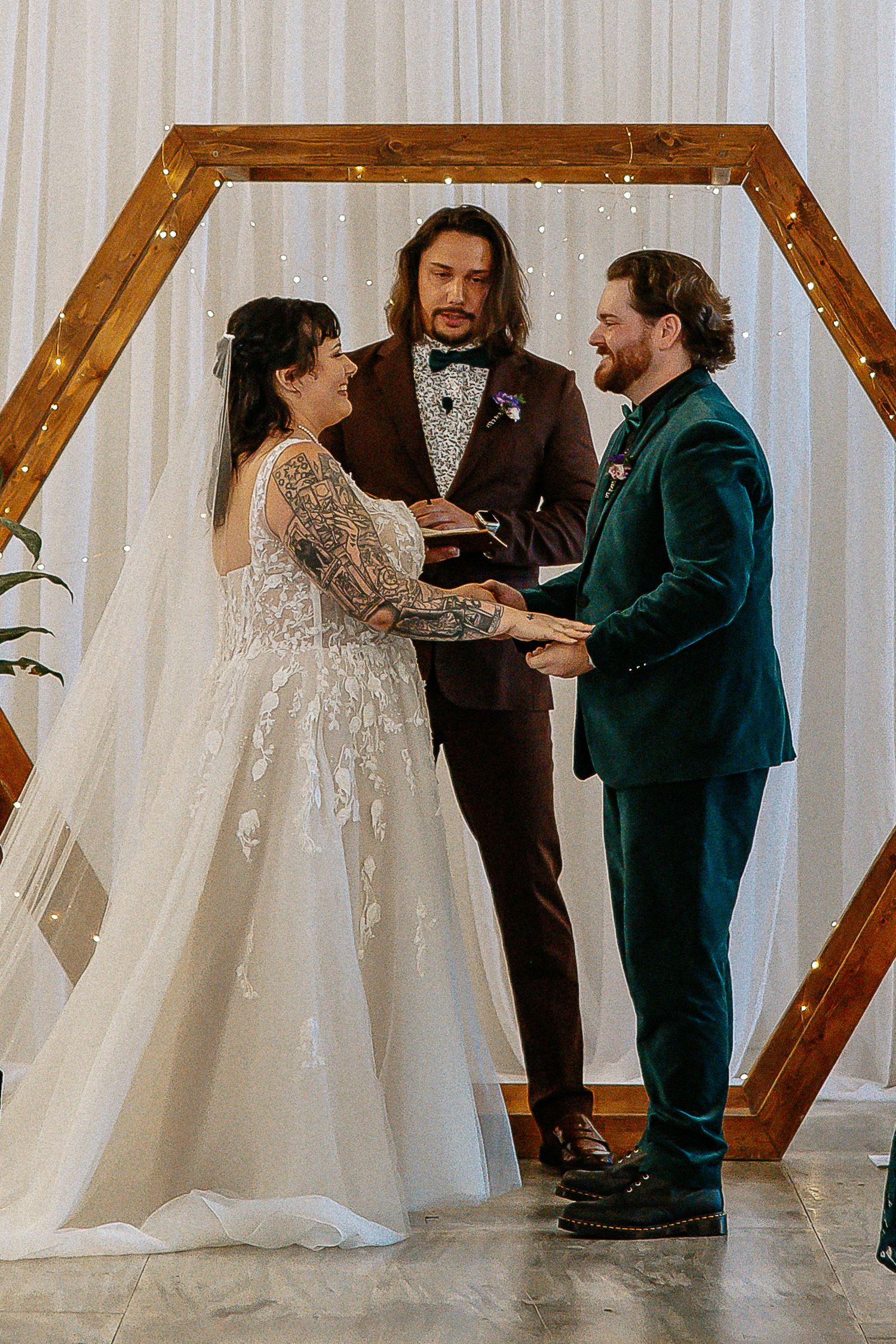 Bride and groom hold hands at altar under wooden hexagon arch with officiant in brown suit.