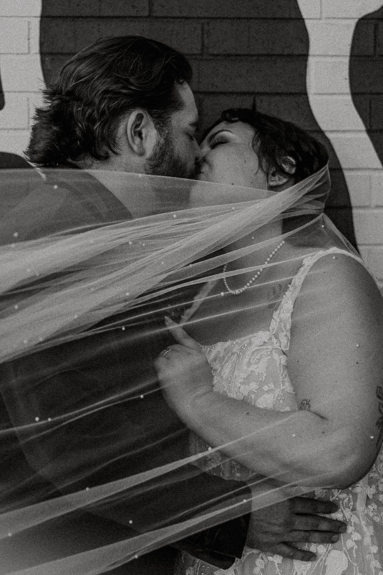 Black and white photo of bride and groom sharing first kiss under flowing sparkle-adorned wedding veil.