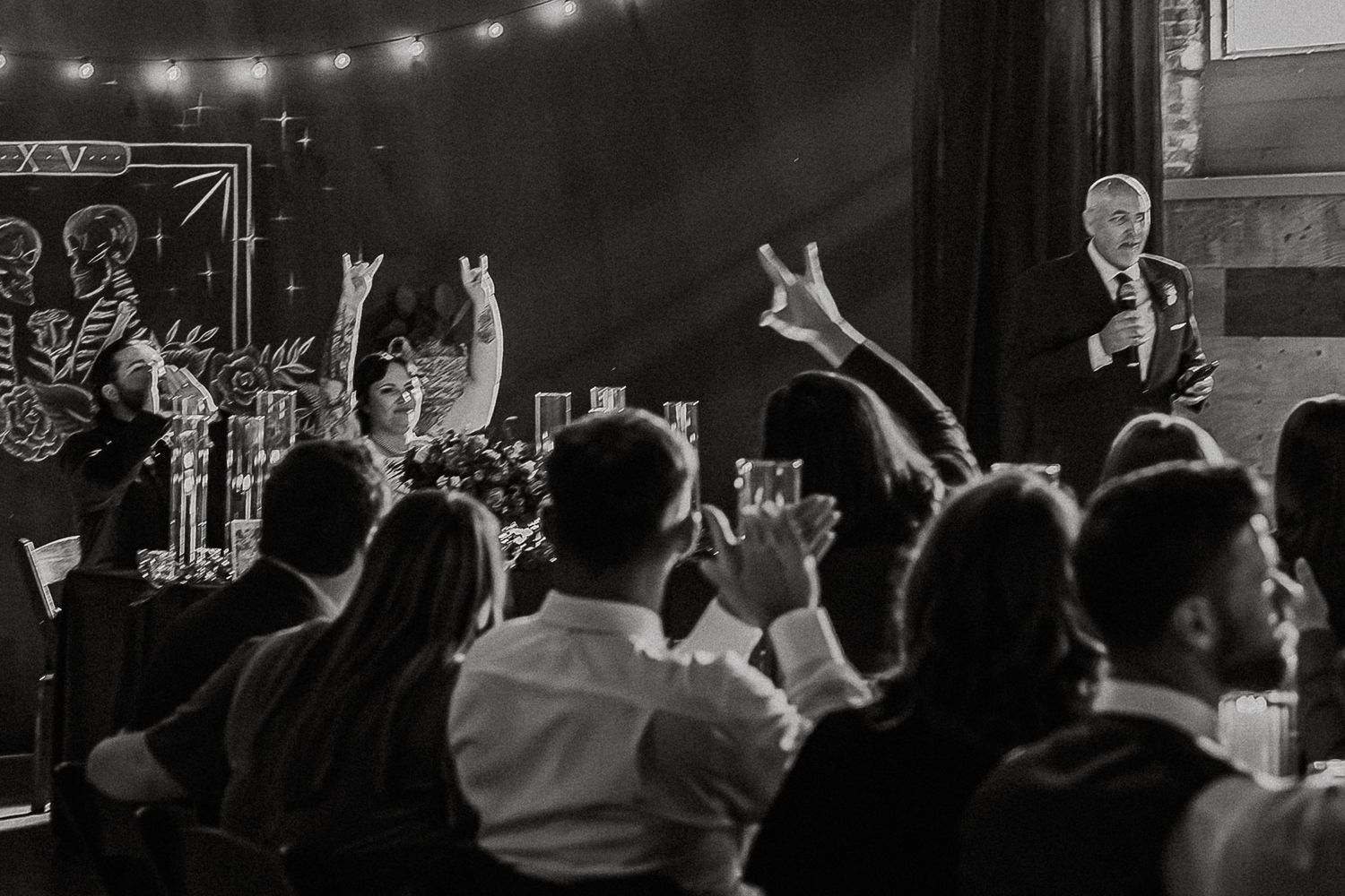 Black and white photo of wedding guests cheering and raising hands during a lively reception toast.