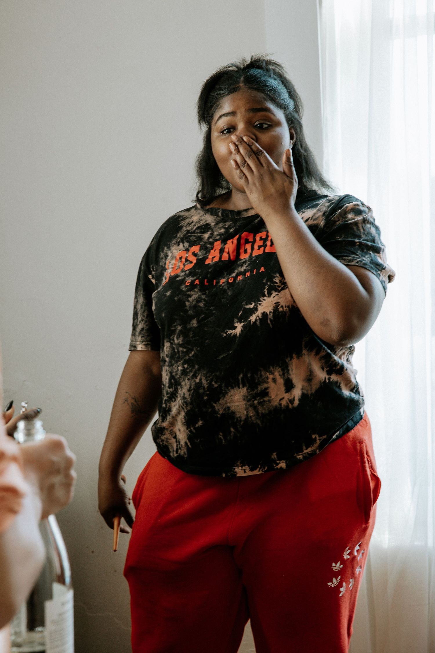 A woman in a tie-dye Los Angeles shirt and red pants covers her mouth in surprise near a bright window at Urban Artifact in Cincinnati, Ohio