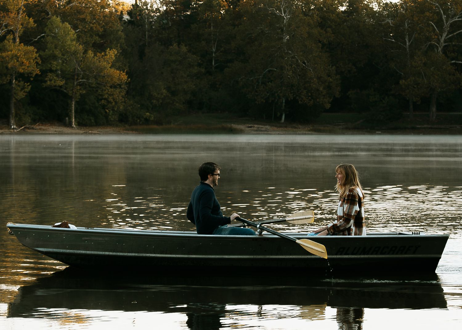 A couple enjoys a peaceful rowboat ride together on a calm, tree-lined lake in autumn in Cincinnati, Ohio