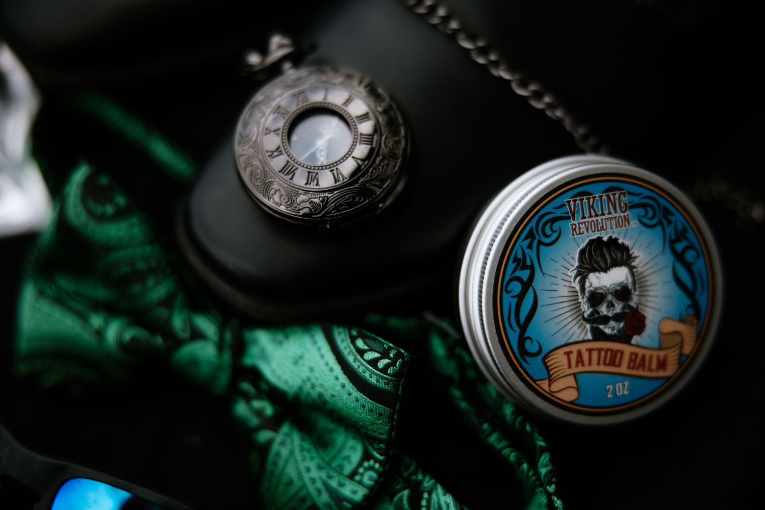Close-up of tattoo balm tin and vintage pocket watch on dark leather surface with green fabric in Cincinnati, Ohio.