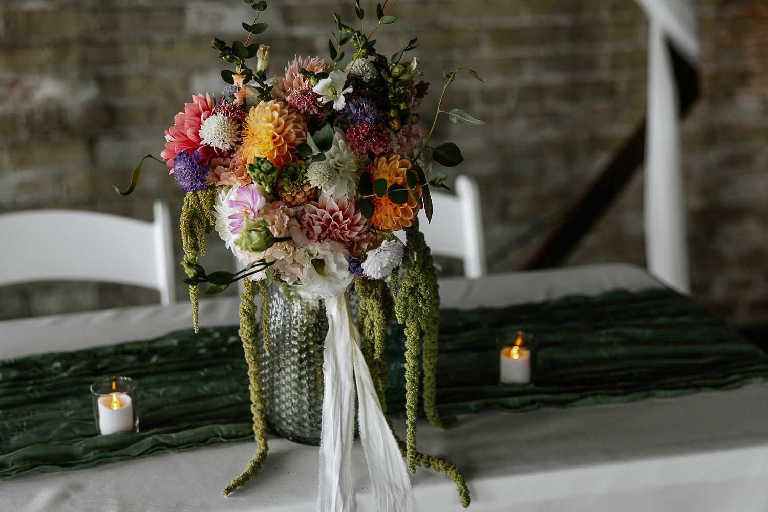 Colorful wedding centerpiece bouquet with dahlias and greenery on white table with candles in rustic venue at The Remedy in Dayton, Ohio