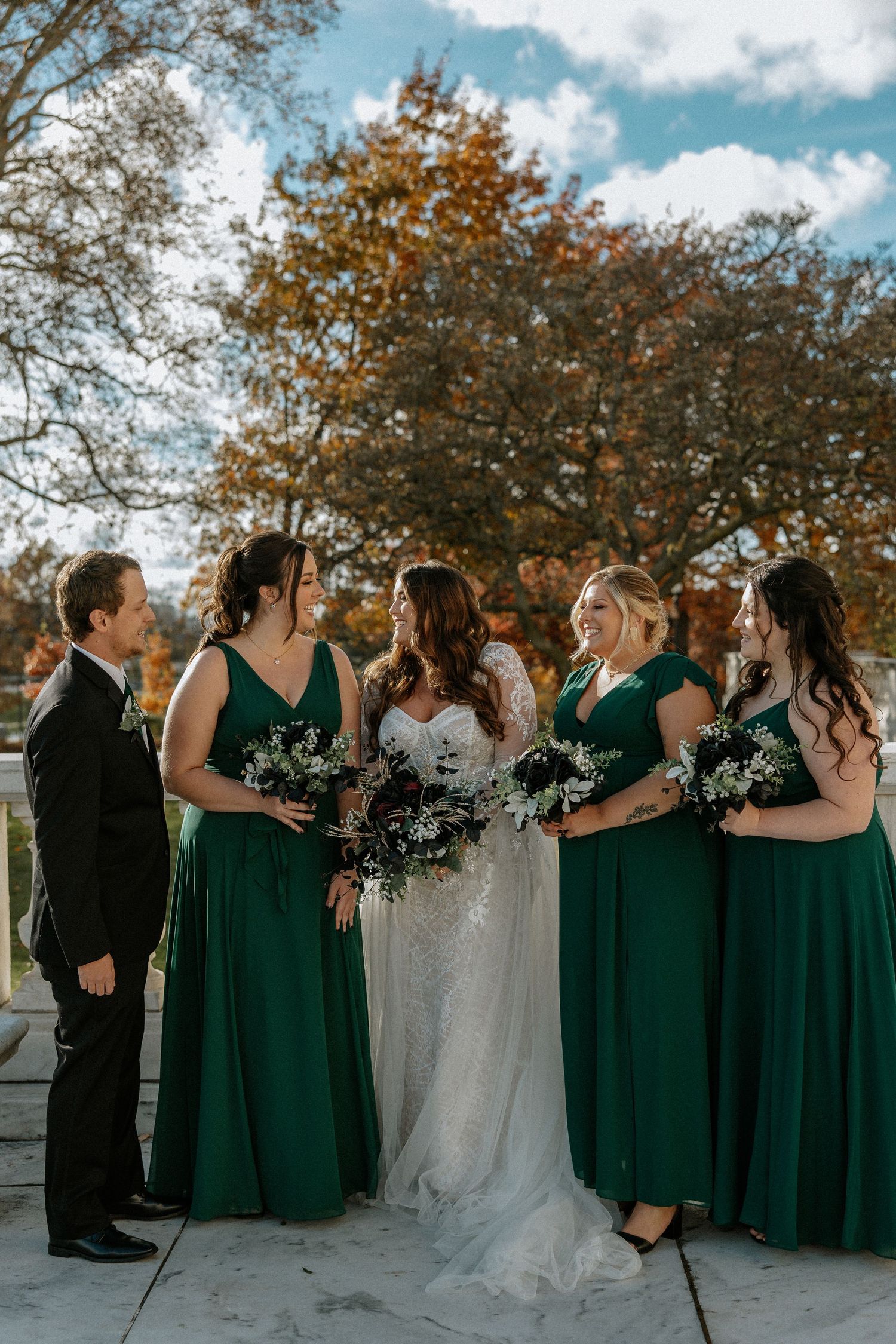 A bride in white lace stands with bridesmaids in emerald green gowns holding bouquets outdoors at The Red Space in Cleveland, Ohio
