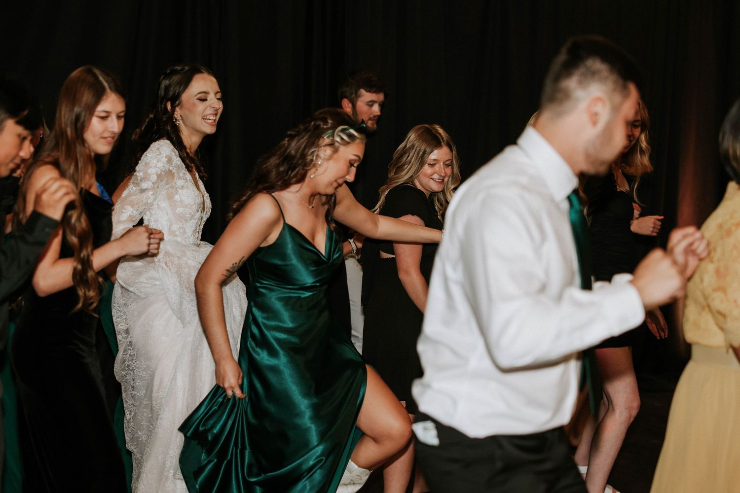 Bride in white lace gown dances with bridesmaids in emerald green velvet dresses at a lively wedding reception.