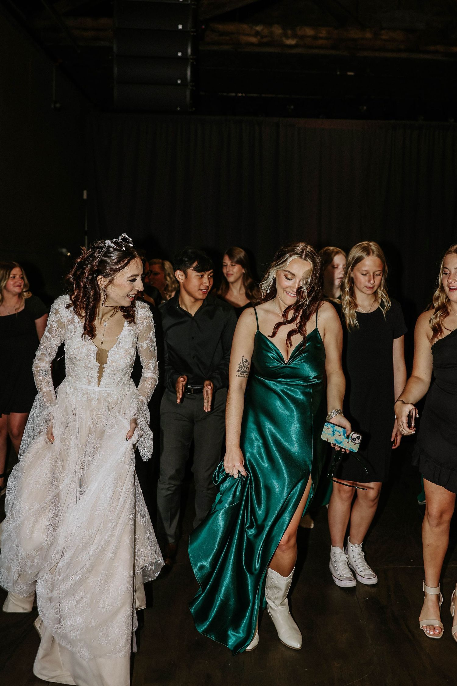 Bride in white lace gown dances with guests in emerald satin dress at wedding reception.