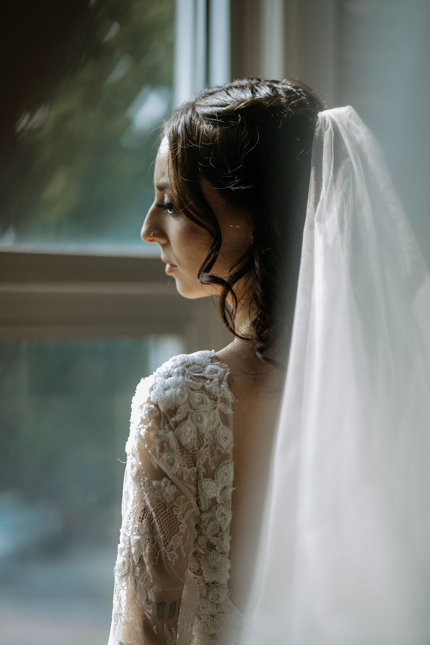 Bride in lace wedding dress and veil gazing thoughtfully out a window in soft natural light.
