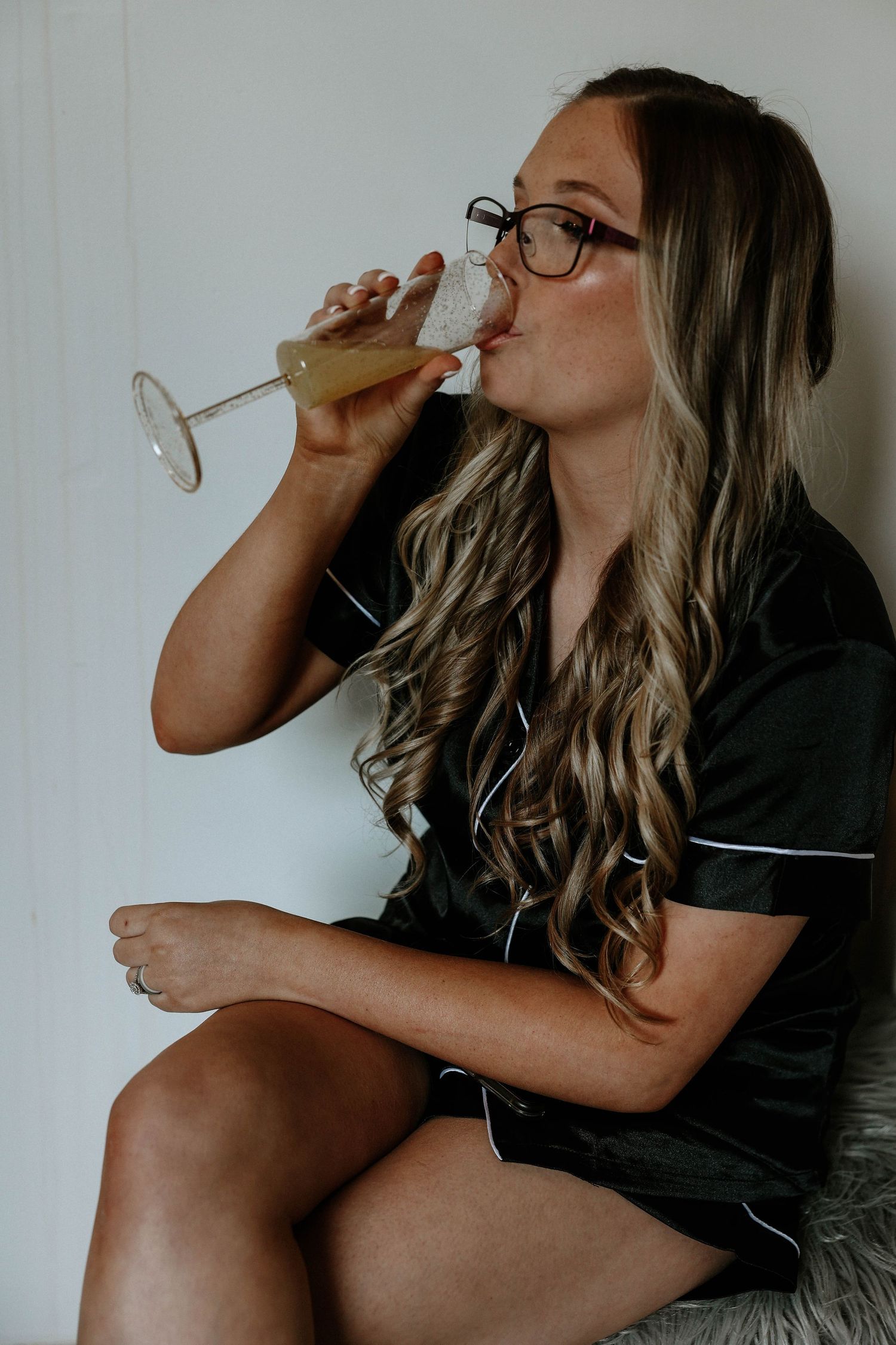 Woman in black satin pajamas drinking champagne from a flute while seated, wearing glasses with long wavy hair.