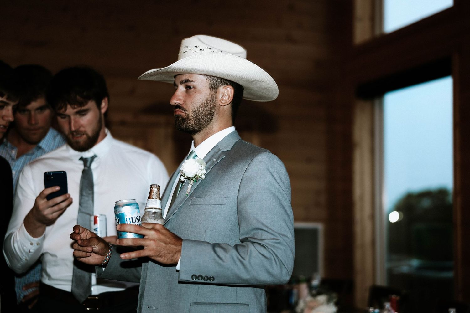 Stylish groomsman in gray suit and white cowboy hat holds drinks at a rustic wedding reception.