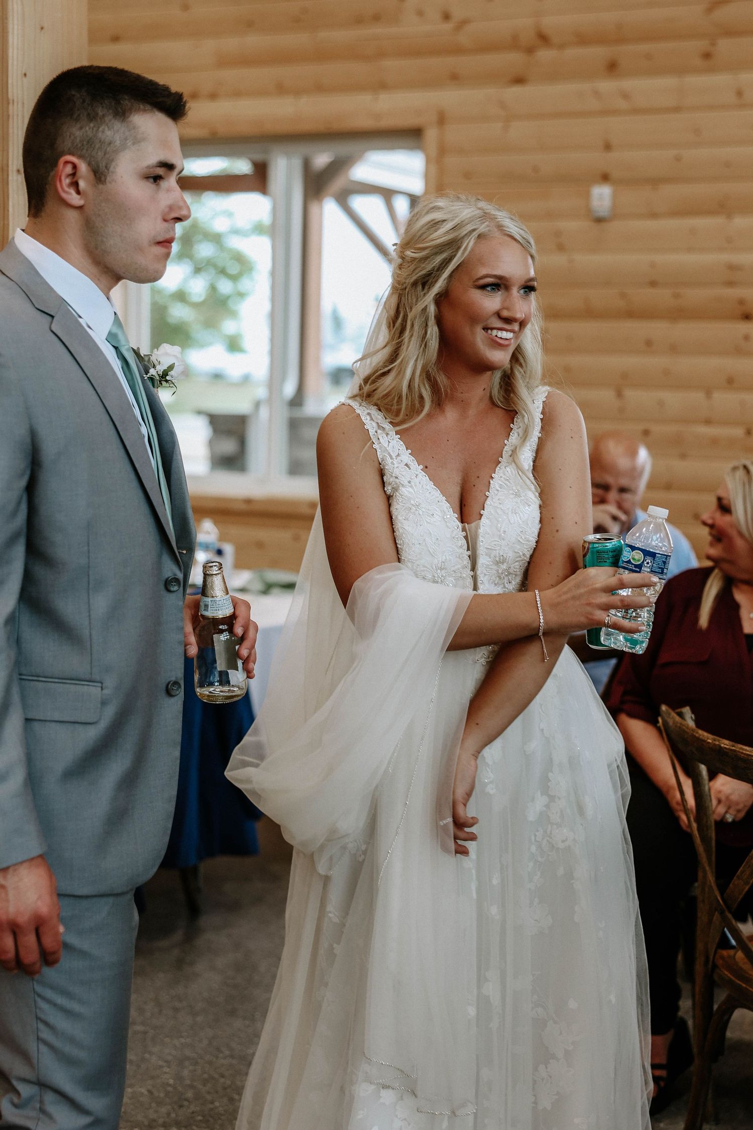 Smiling bride in white lace gown holding water bottle stands beside groom in gray suit at rustic barn wedding reception.