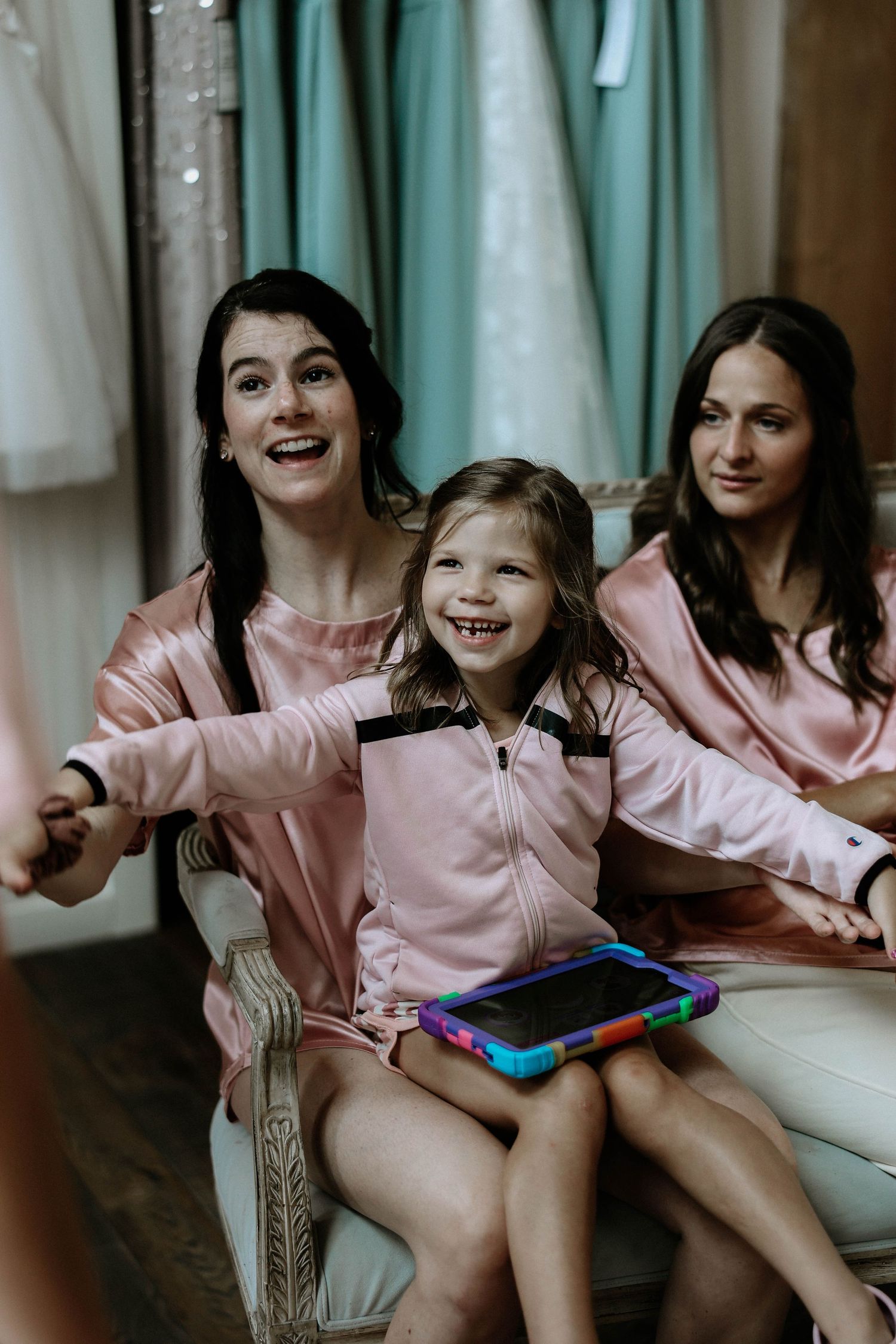 Bridesmaids in pink satin robes laughing with a young flower girl holding a tablet before the wedding.