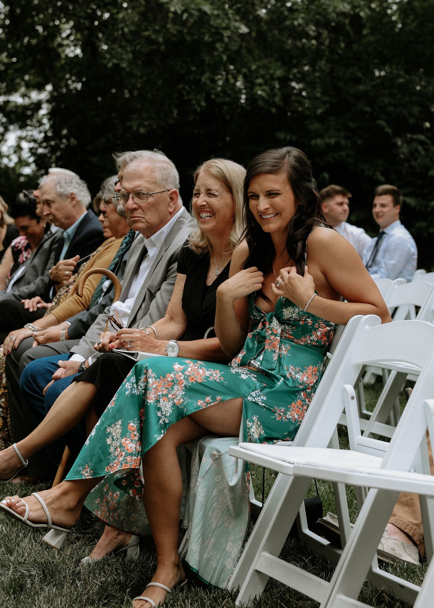 Smiling wedding guests seated in white chairs at an outdoor ceremony, wearing floral dresses and formal attire.