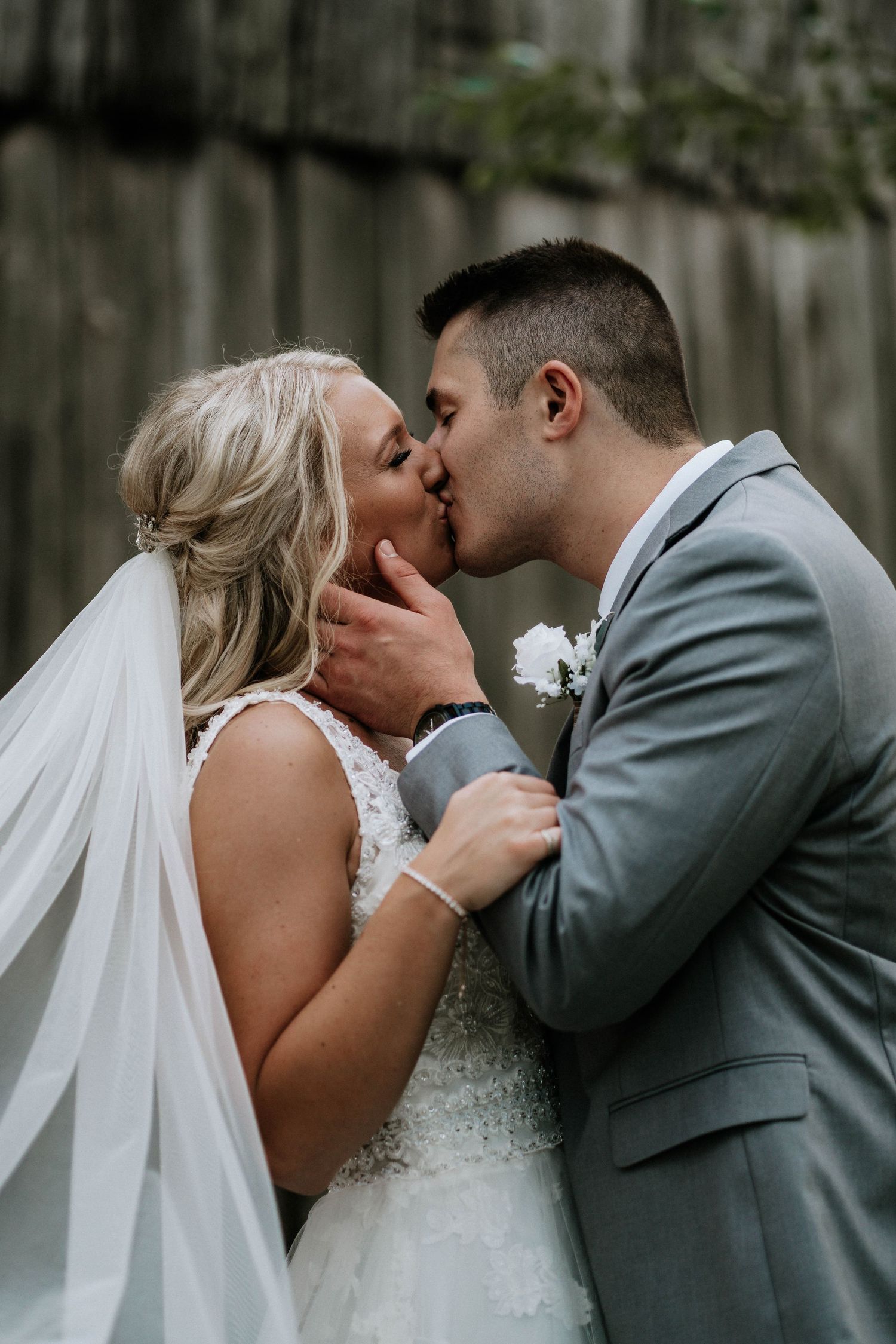Bride and groom share a romantic kiss, bride in lace gown with veil, groom in gray suit with white boutonniere.