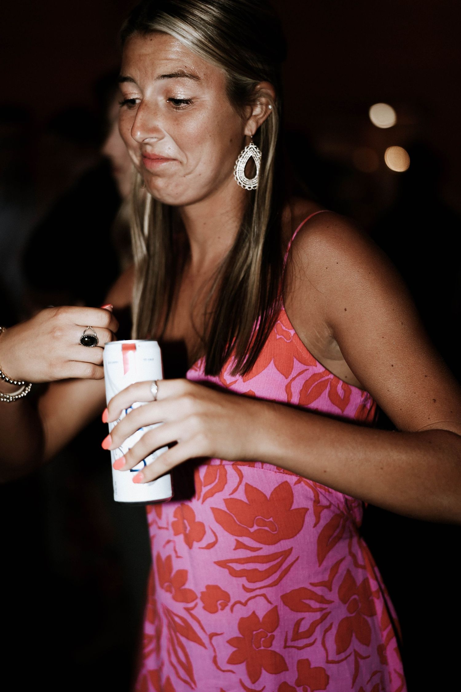 Woman in pink floral dress sipping a drink through a straw at a party, wearing drop earrings.