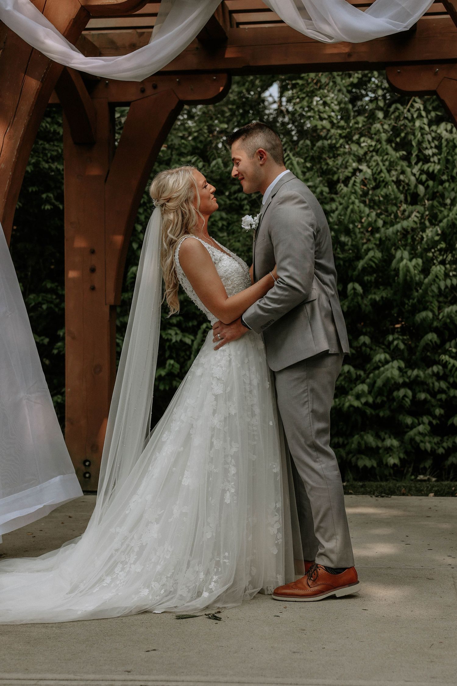 Bride and groom share a romantic moment under a wooden pergola draped with white fabric at an outdoor wedding.