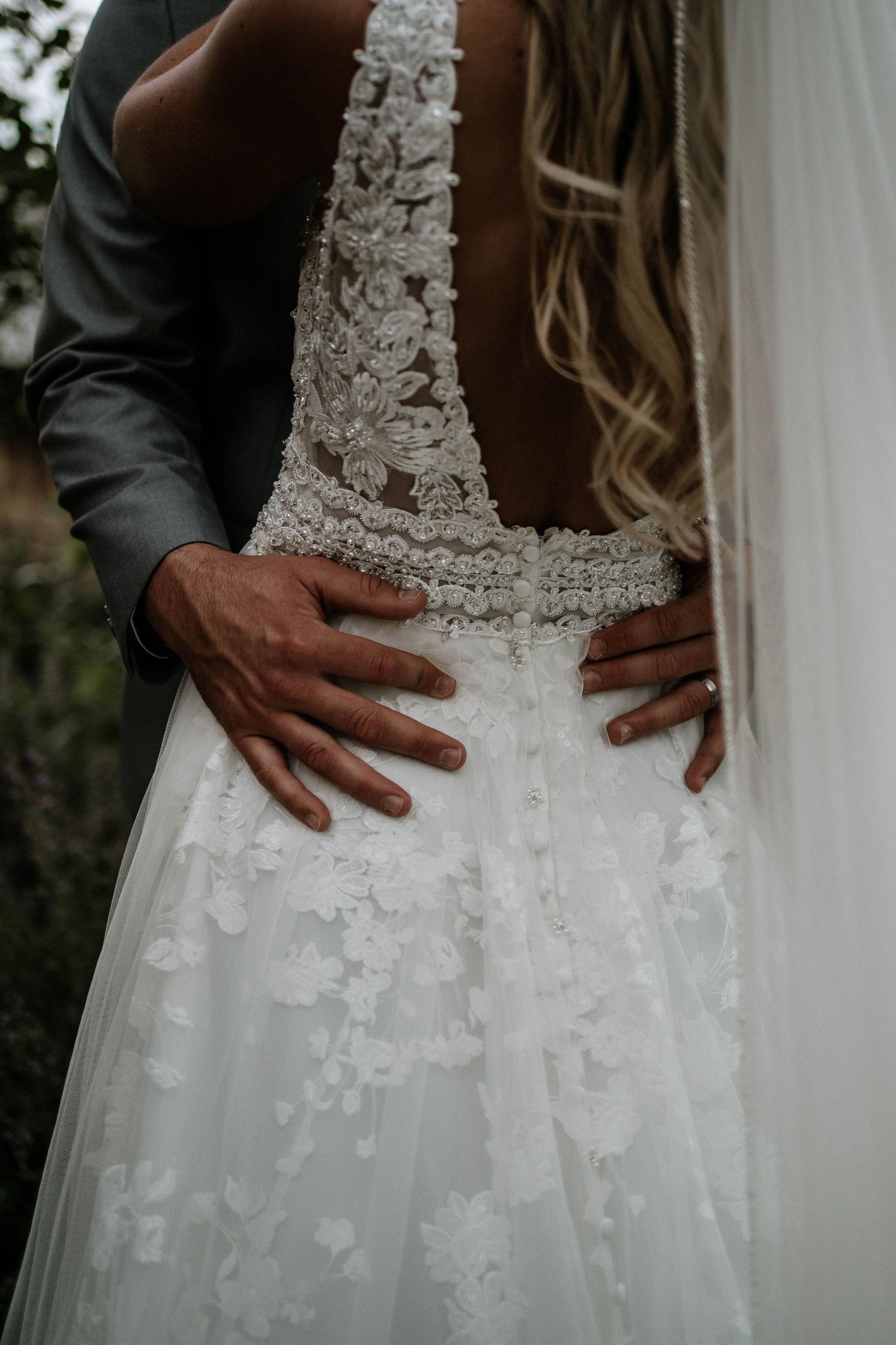 Groom's hands on bride's waist, showcasing intricate lace back wedding gown with floral appliques and flowing tulle skirt.