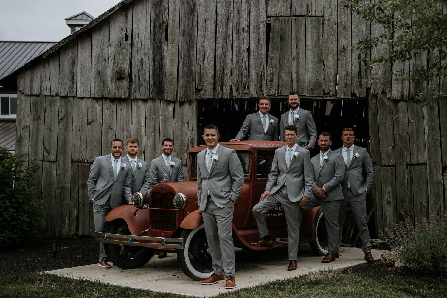 Groomsmen in gray suits posing with vintage rustic red car in front of wooden barn.