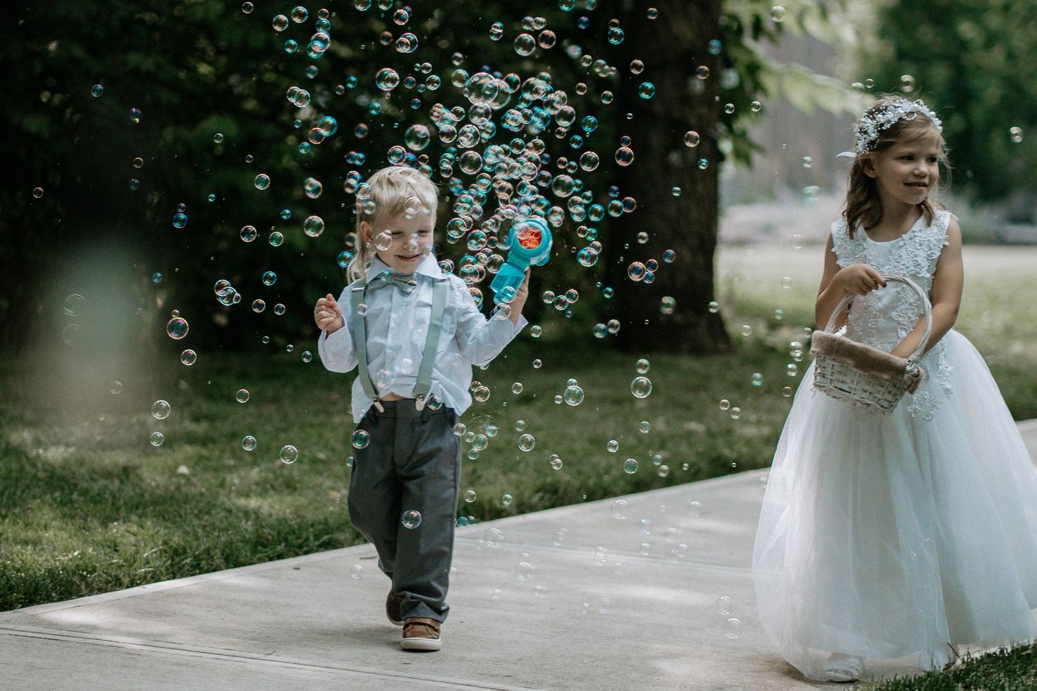 Children walking down wedding aisle, boy blowing bubbles with toy gun, flower girl in white dress smiling.