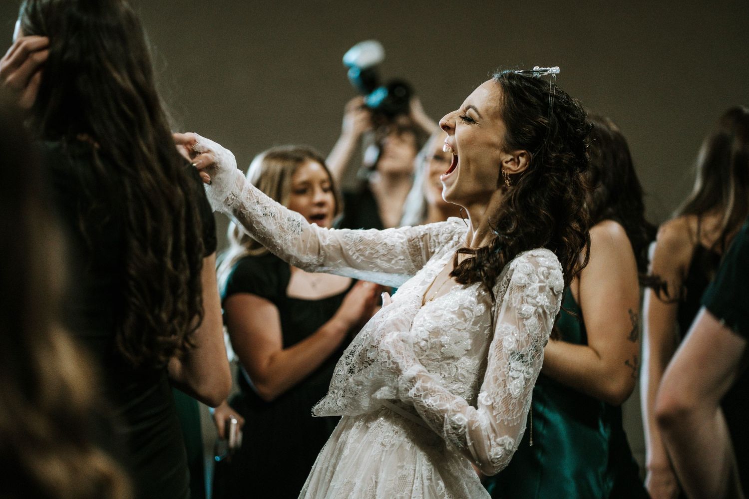 Joyful bride in lace gown laughing and dancing with bridesmaids in emerald dresses at wedding reception.