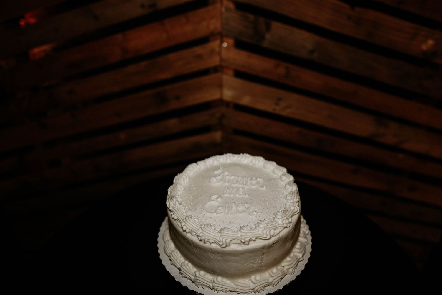 White two-tier wedding cake with elegant piped frosting details displayed on a table in a rustic wood-paneled venue.