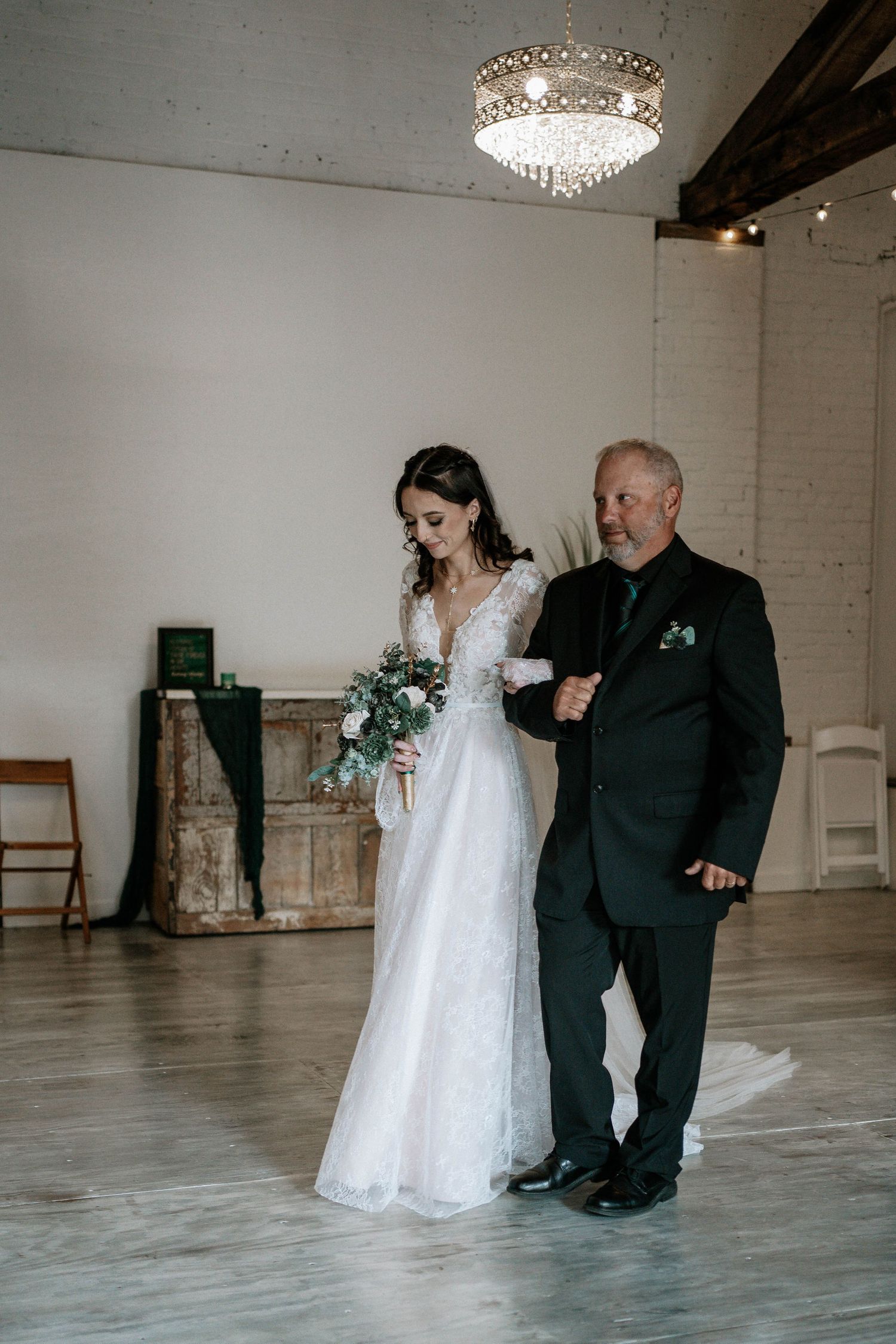 Bride in lace gown holding bouquet walks down aisle with escort in black suit under crystal chandelier.