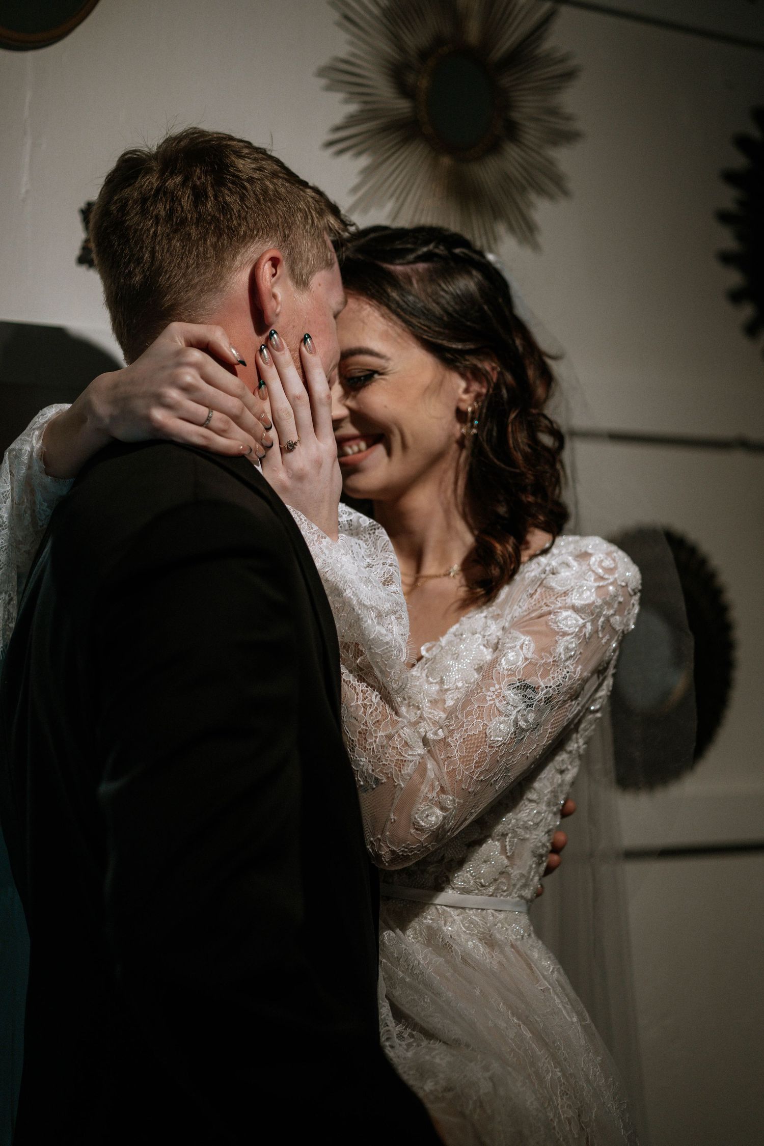 Bride in lace gown smiles while holding groom's face during romantic wedding moment indoors.