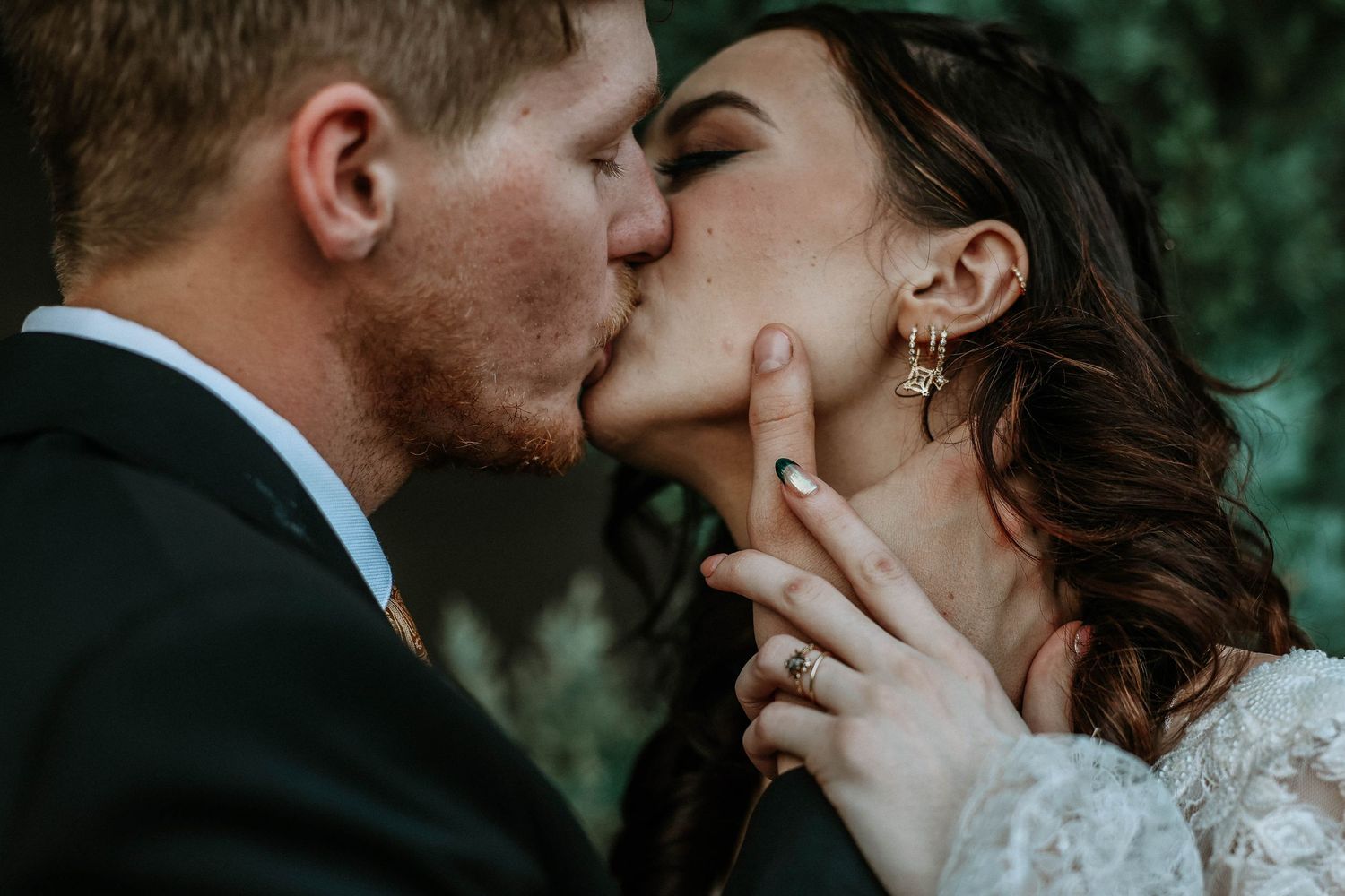 A bride and groom share a romantic kiss on their wedding day, with the bride wearing a lace dress and gold earrings.