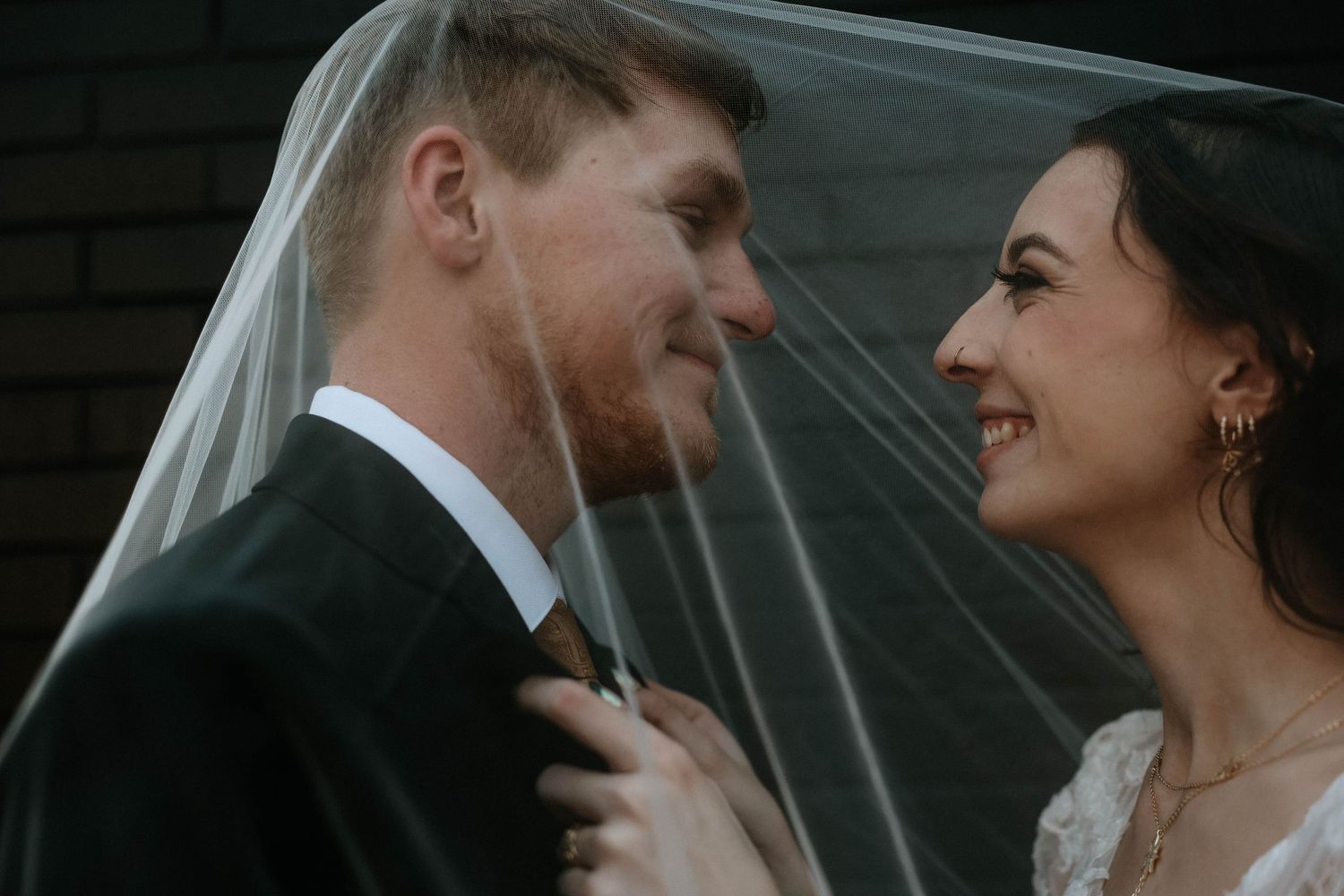 Bride and groom share an intimate moment under a wedding veil, smiling at each other on their wedding day.