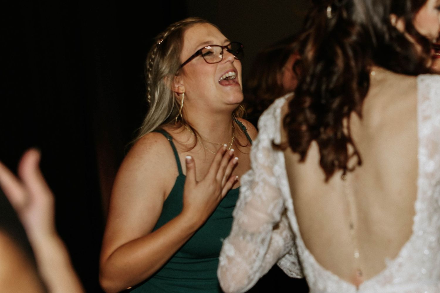 Wedding guest in green dress and glasses laughing joyfully near bride in white lace gown at reception.