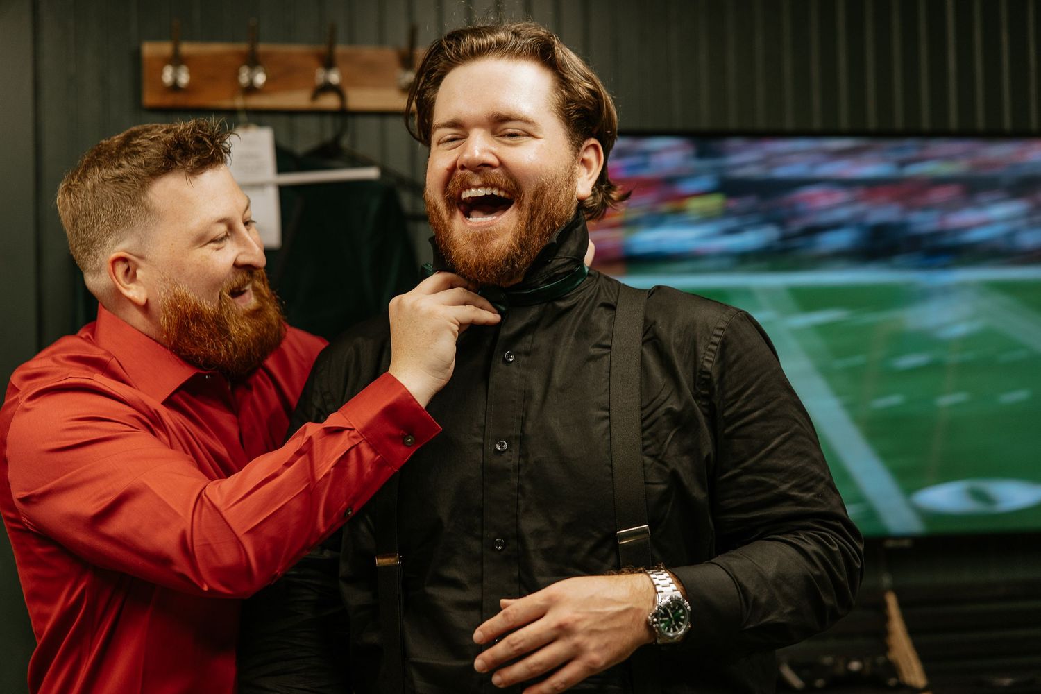 Two bearded men laughing while one adjusts the others collar before a wedding ceremony.