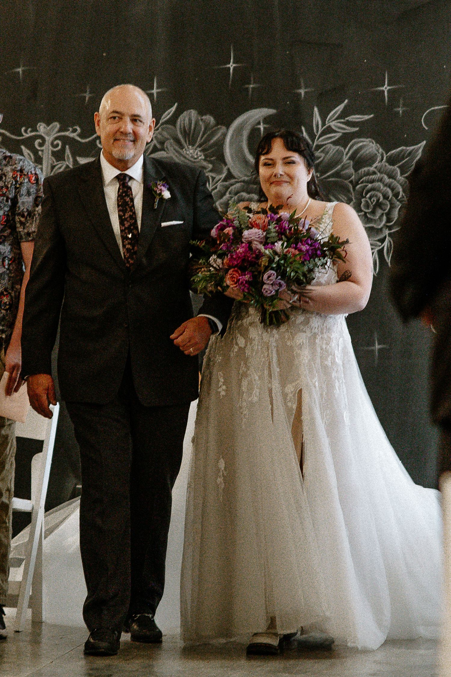 Bride in lace gown holds colorful bouquet while walking down aisle with father.
